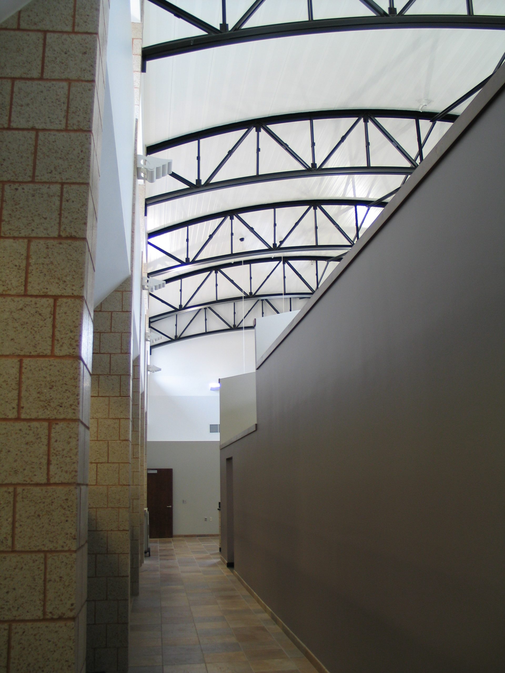 A hallway with tile flooring, tan brick walls on the left, and a high ceiling supported by black metal beams. Natural light shines in from upper windows, creating a bright, open atmosphere.