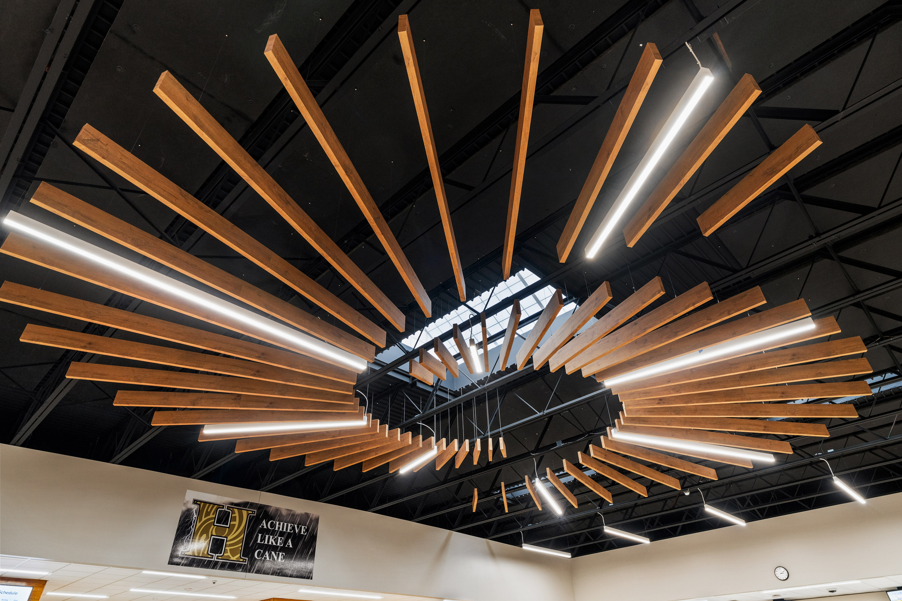 A modern ceiling installation features radiating wooden beams and linear LED lights arranged in a circular pattern, with a black metal ceiling and exposed beams visible above.
