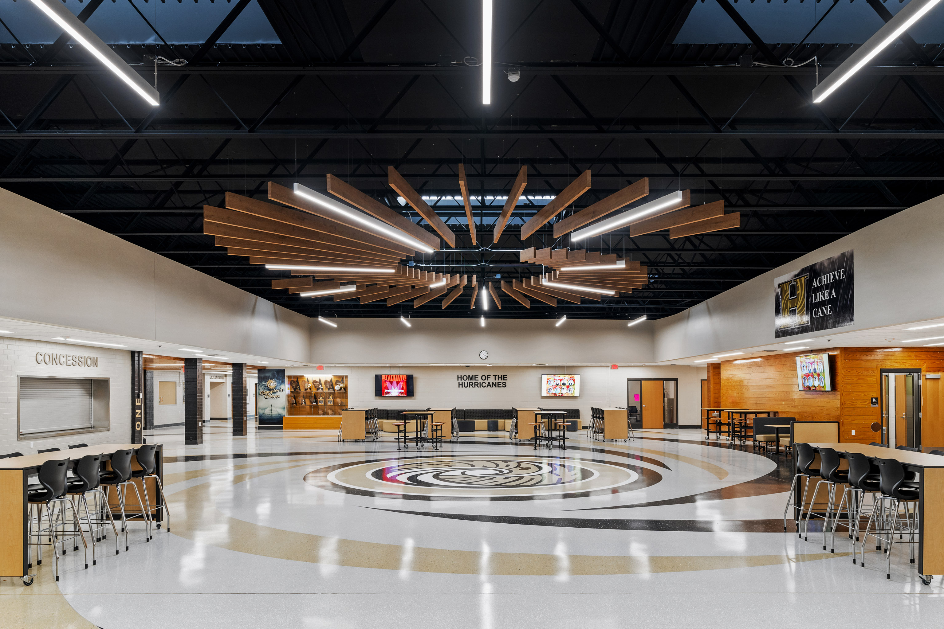 A spacious, modern school cafeteria with circular patterns on the floor, black and wood seating, and a dramatic wooden chandelier under a skylight. Walls have concession stands, a trophy case, and school banners.