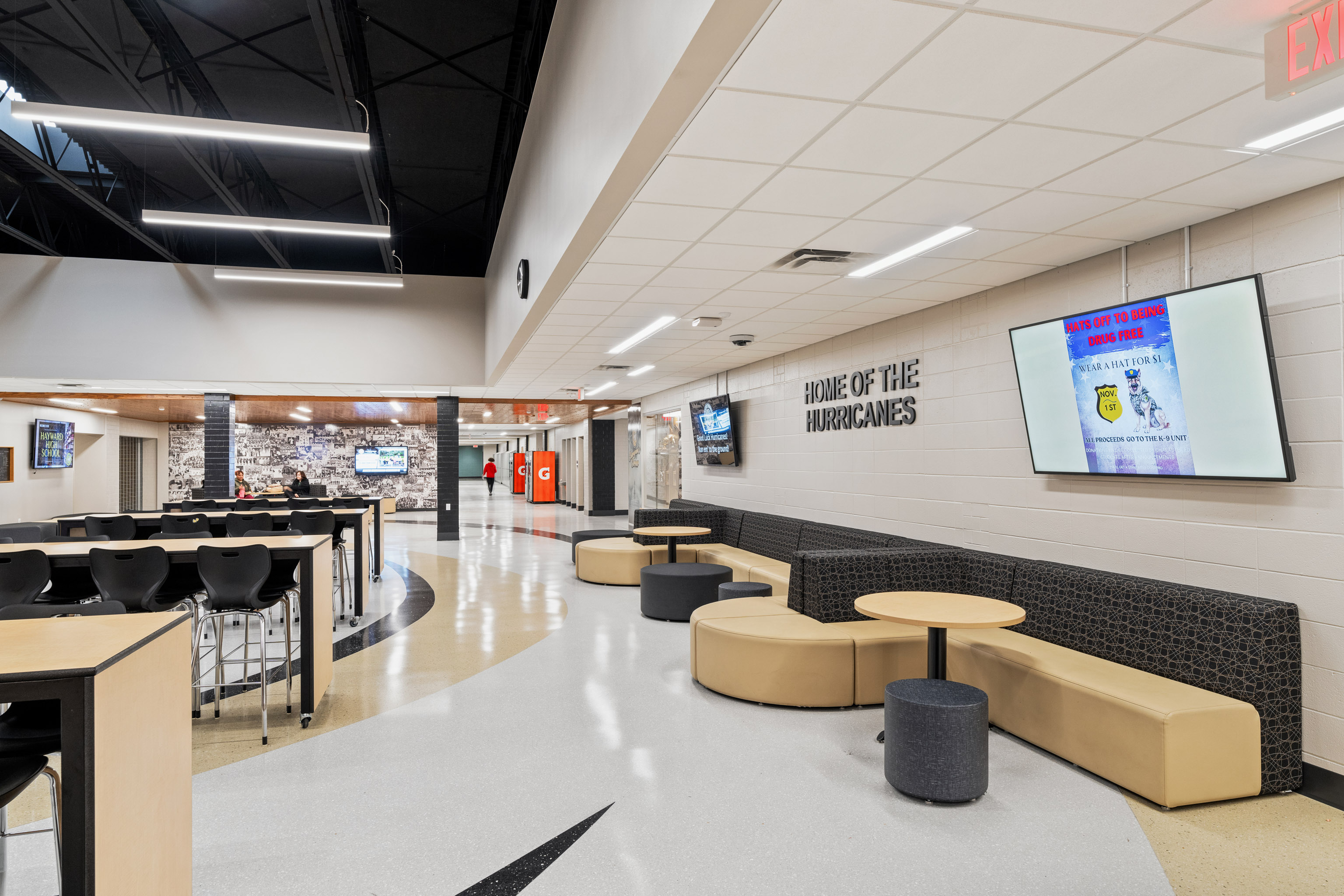 A modern school common area with curved beige seating, round tables, black chairs, and wall signs reading "HOME OF THE HURRICANES." Digital screens and bright lights are visible, with hallways in the background.