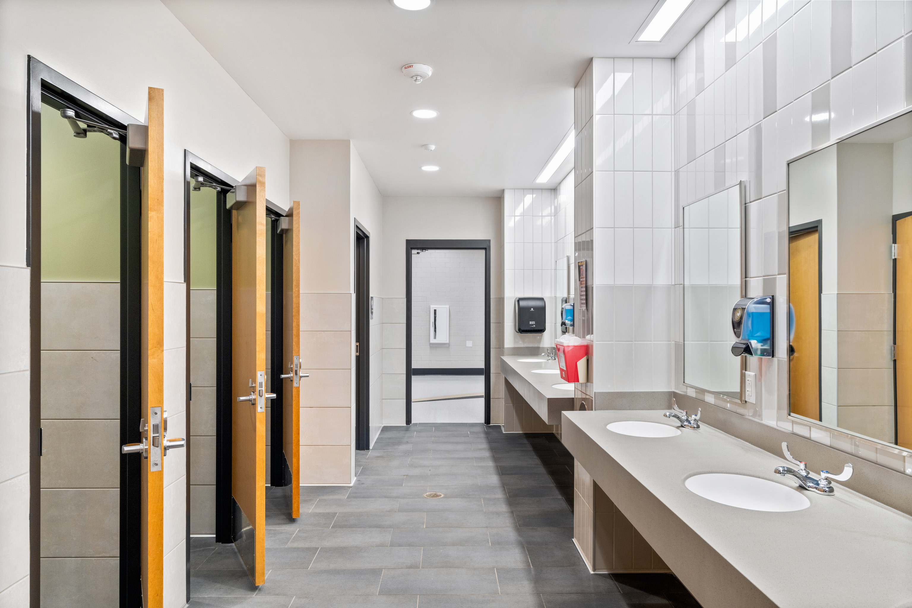 A clean, modern public restroom with three stalls, two sinks, a soap dispenser, mirrors, and a baby changing station. The space has tiled walls, gray flooring, and bright overhead lighting.