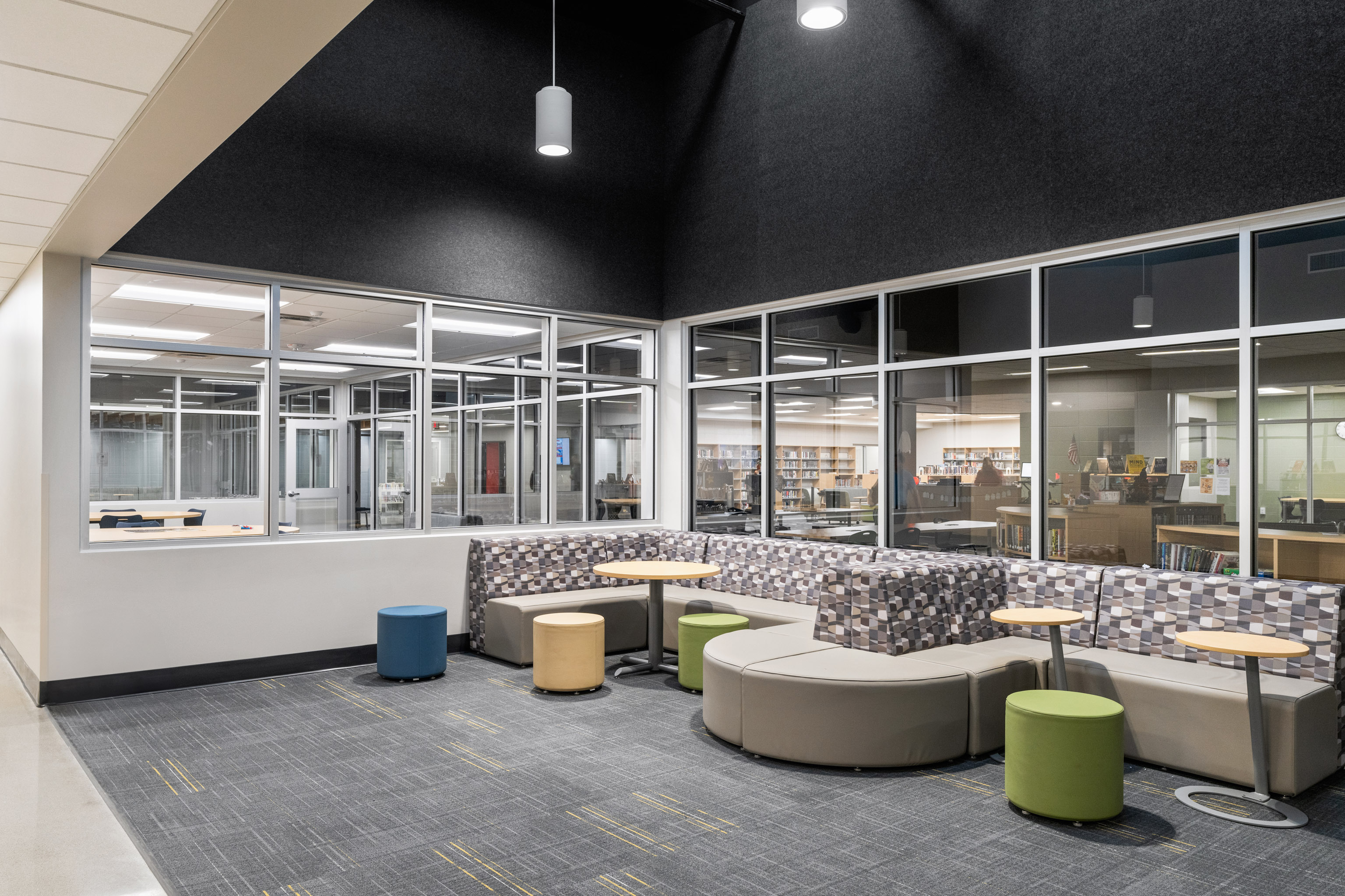 A modern lounge area with patterned and curved seating, round stools, and small tables, set near large glass windows overlooking a library space. The room is well-lit with a gray carpeted floor.