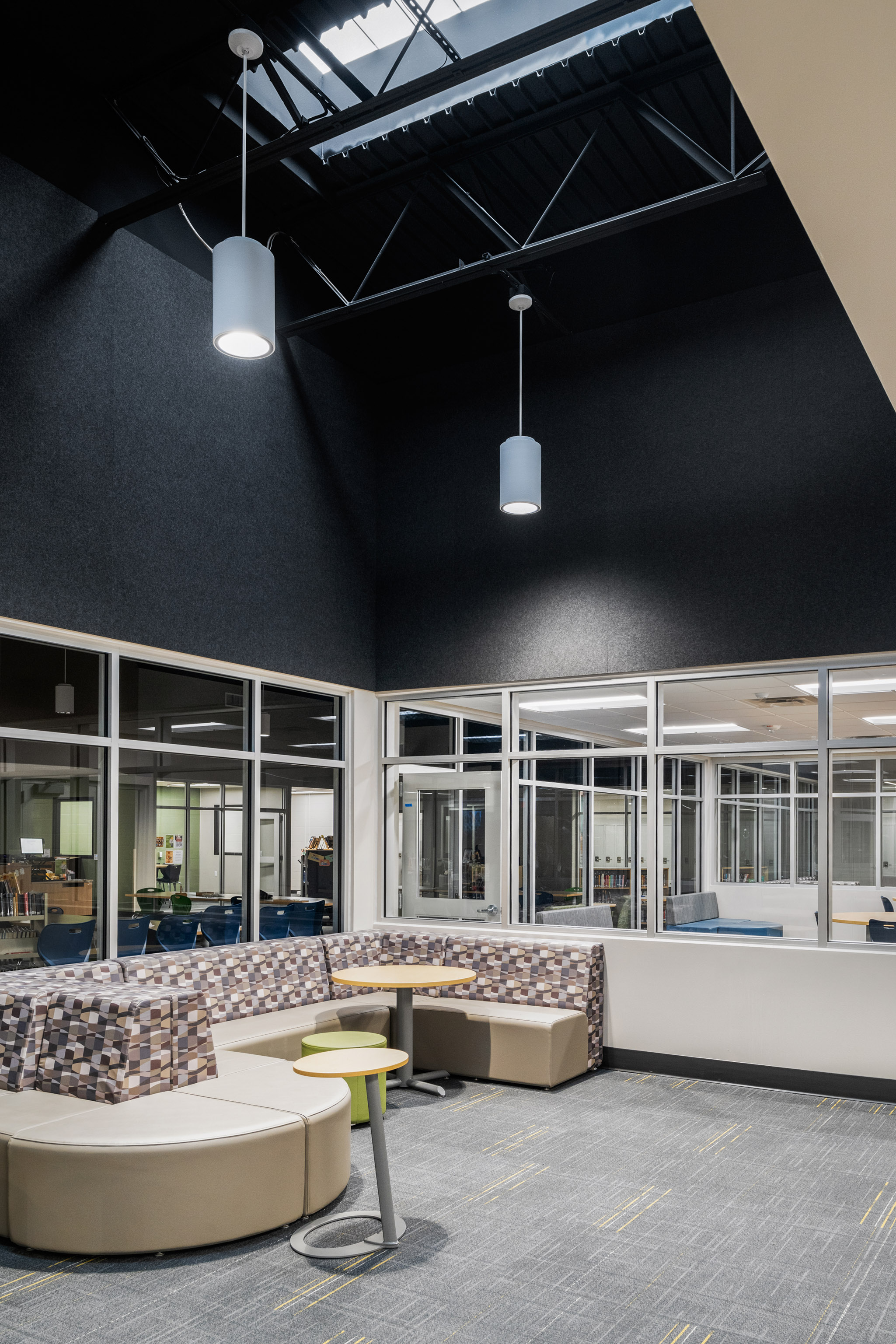 Modern lounge area with patterned and beige seating, small tables, and large windows, under high ceilings with hanging lights and exposed beams. Natural light enters through a skylight above.