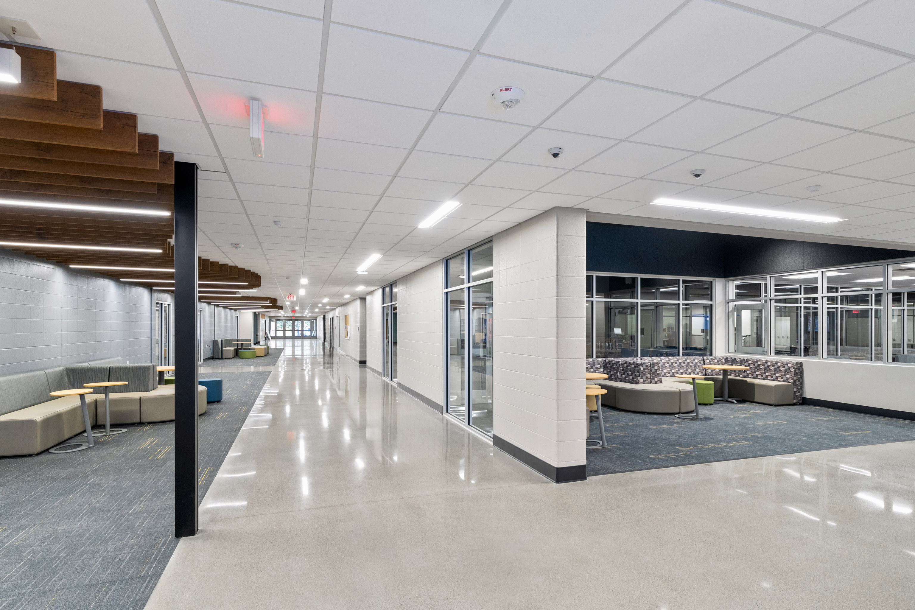 A bright, modern school hallway with polished floors, white walls, and glass-walled seating areas featuring chairs and tables. Ceiling lights and wood accents add to the contemporary design.