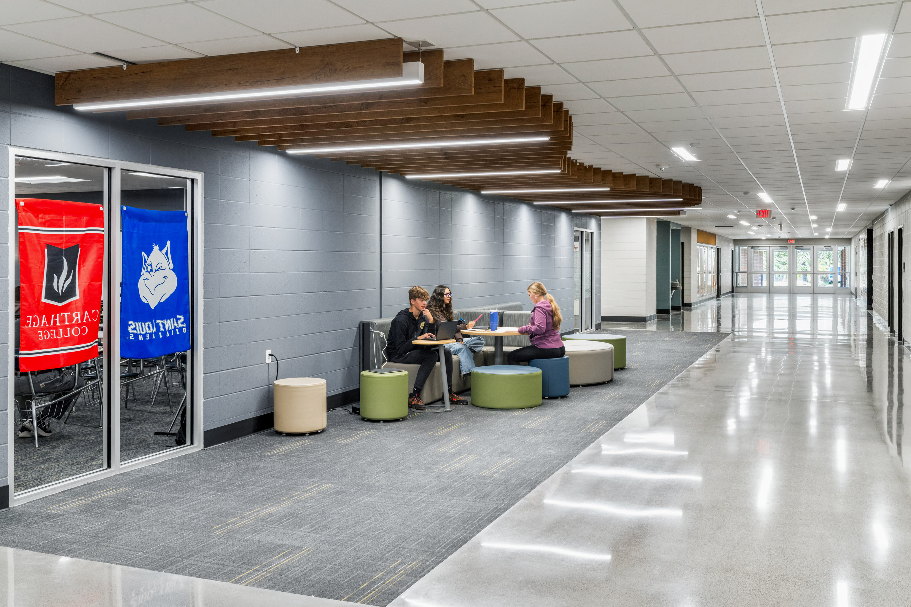 Three people sit and work at a small round table with laptops in a modern school hallway, near glass-walled rooms with college banners. The hallway is bright with gray walls and wood ceiling accents.