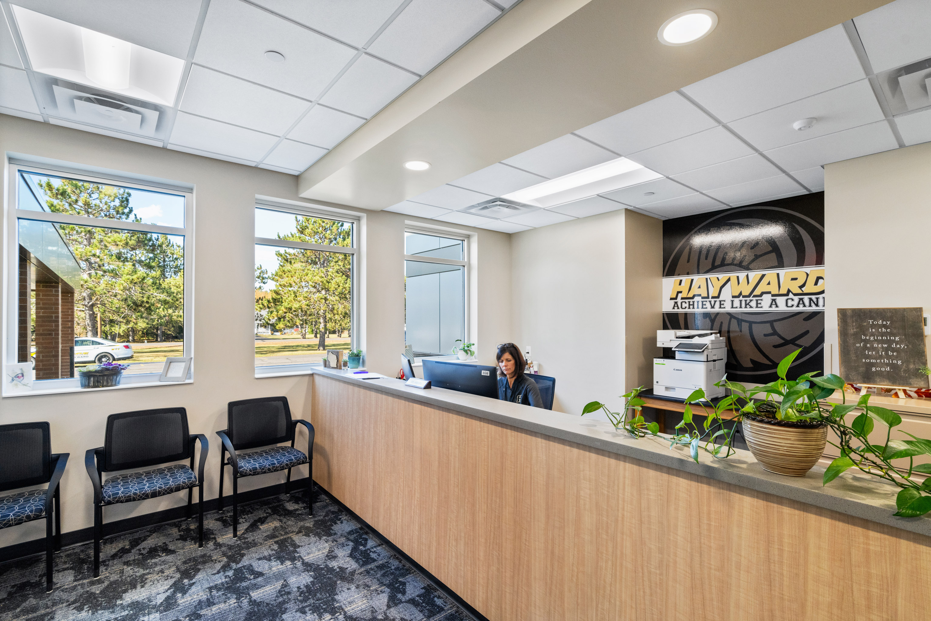 A woman sits at a reception desk in a modern office lobby with several chairs, large windows letting in natural light, and a sign on the wall that reads "HAYWARD ACHIEVE LIKE A CHAMP.