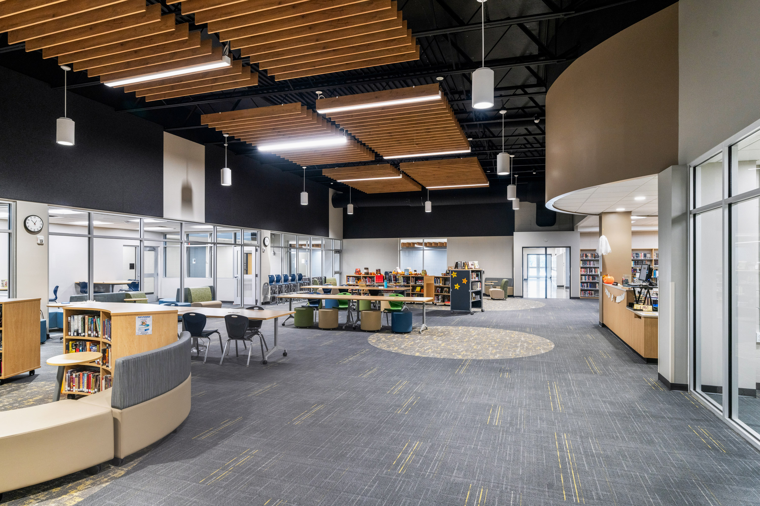 Modern library interior with glass-walled study rooms, bookshelves, round tables, chairs, and curved seating. The ceiling features wooden slats with hanging lights, and the floor is carpeted with a geometric pattern.