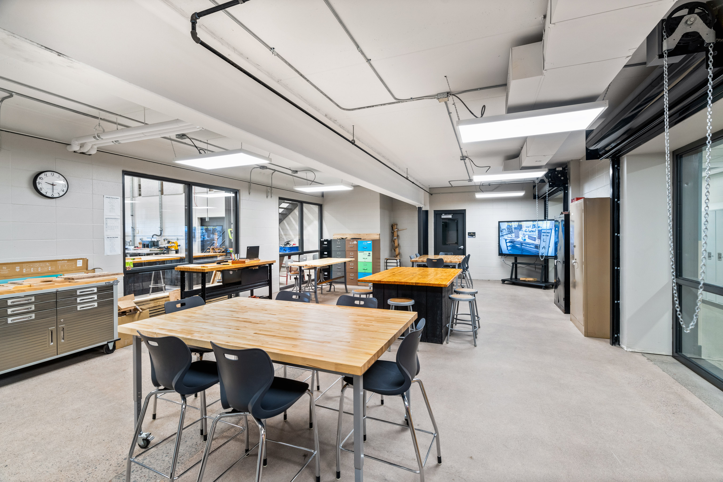 A modern classroom or workshop with wooden tables, black chairs, bright overhead lights, visible exposed pipes, and a wall clock. There are tools, cabinets, and a large TV displaying a workspace.