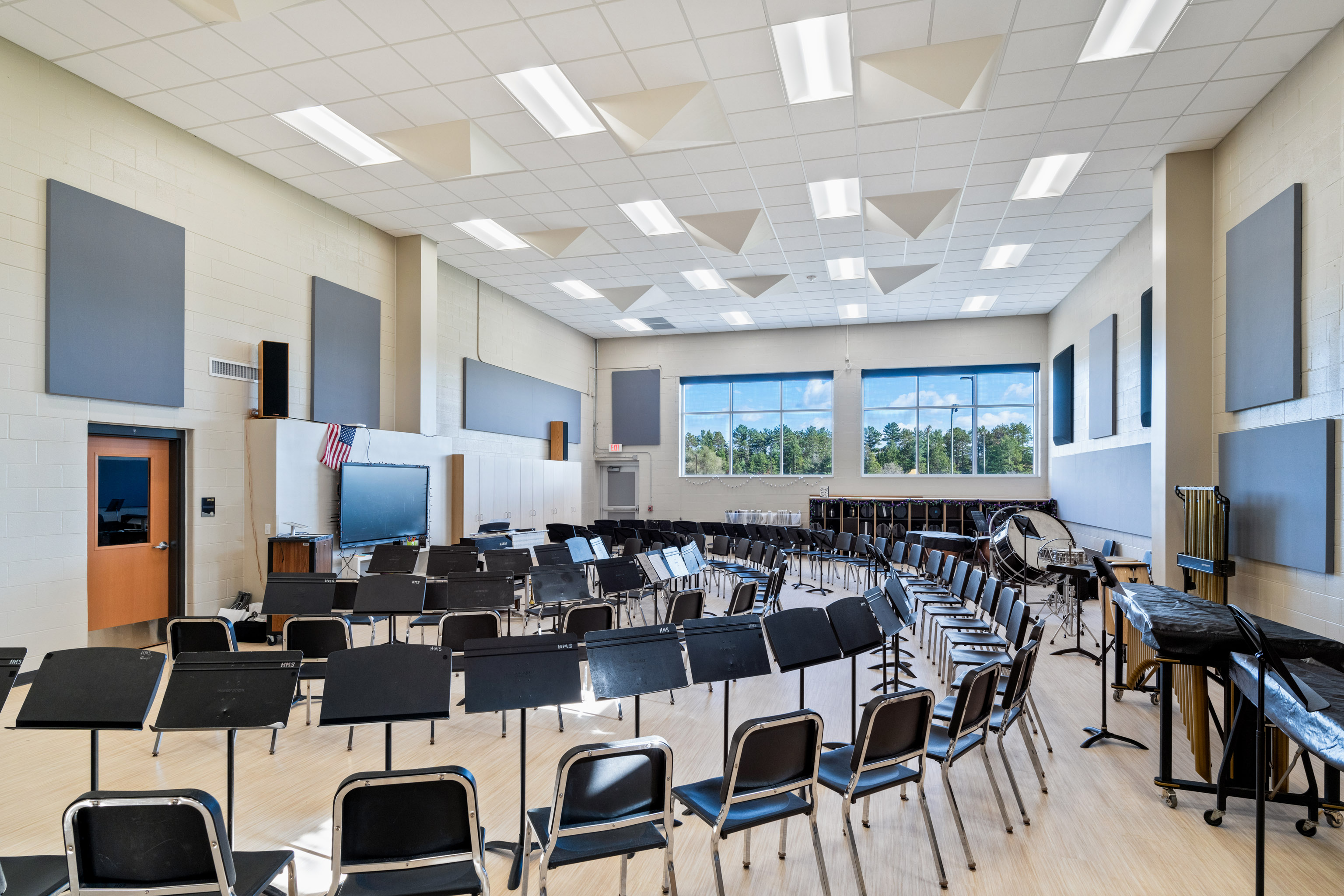 A spacious, well-lit band room with rows of black music stands and chairs arranged in a semicircle. Large windows let in daylight, and percussion instruments are set against the side wall. Acoustic panels cover the walls and ceiling.