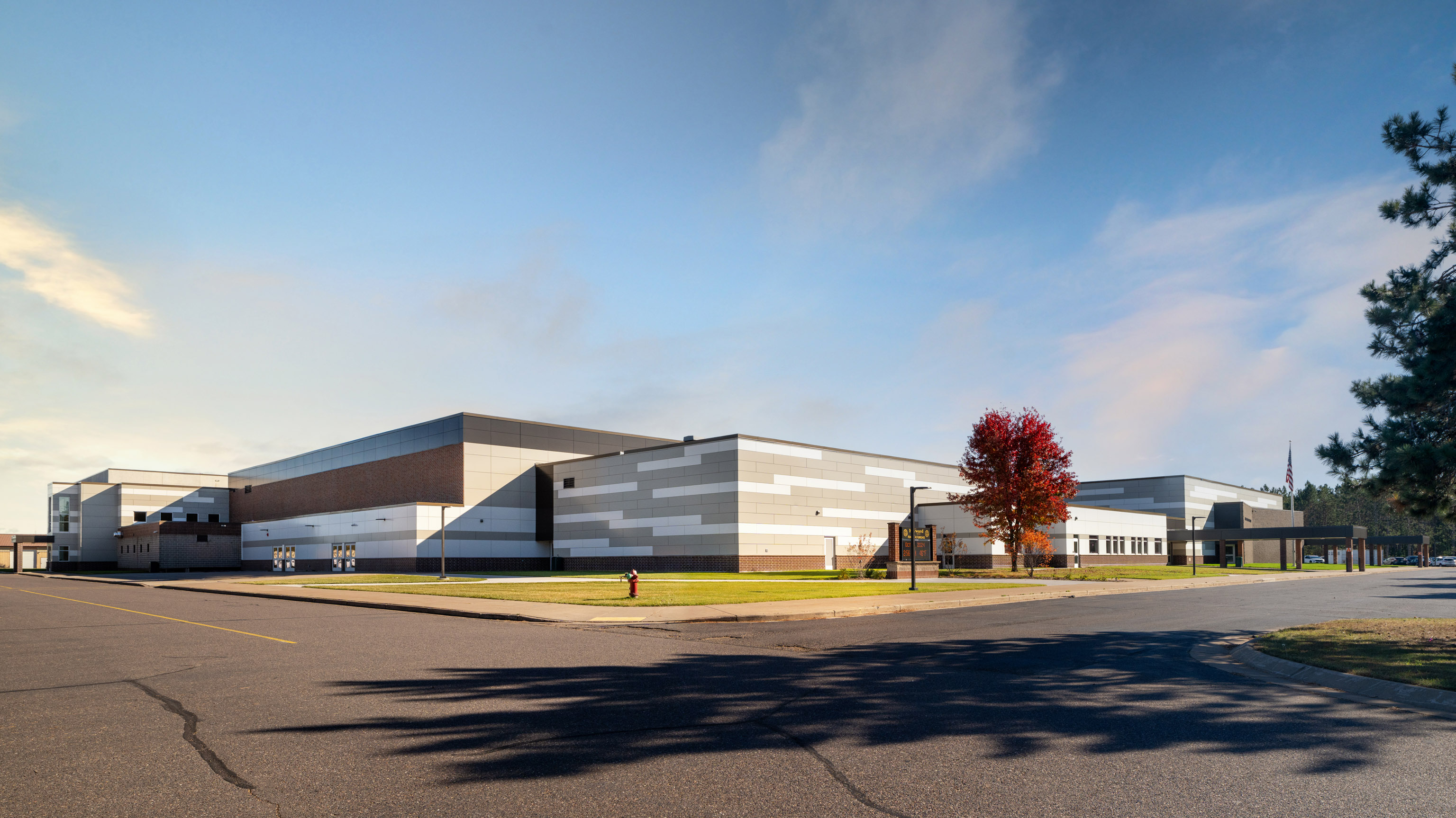 A large modern school building with a mix of gray, white, and brick exterior, surrounded by a paved road and green lawn. A single red-leaved tree is near the entrance under a partly cloudy sky.