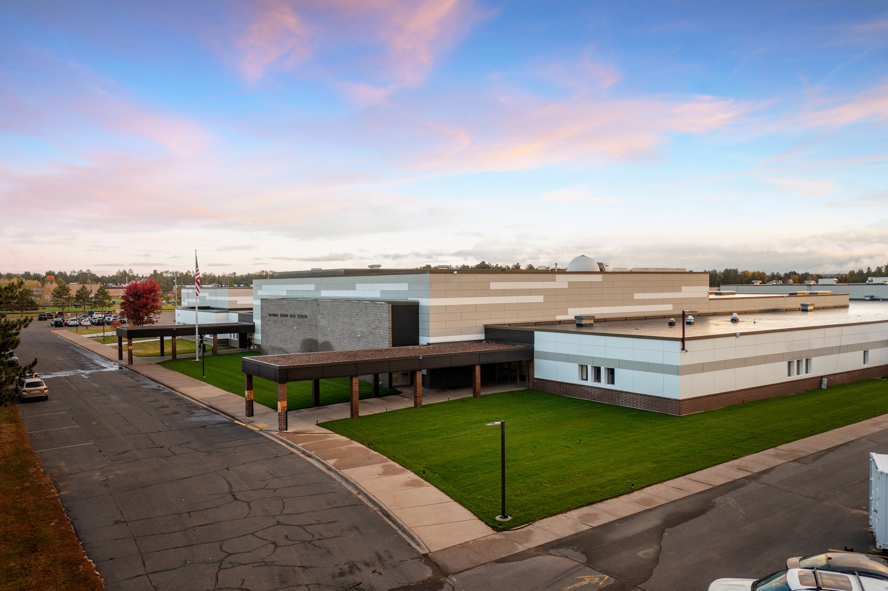 A modern, single-story school building with beige and gray exterior sits next to a street, surrounded by green lawns under a cloudy, colorful sunset sky.