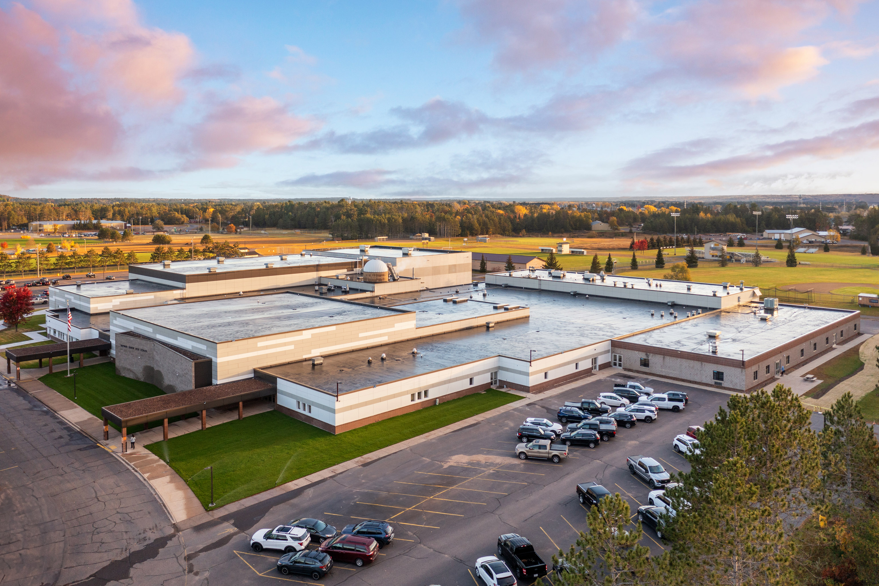 A large, single-story school building with a flat roof sits surrounded by grass and trees. Numerous cars are parked in a lot in front, under a partly cloudy sky with early morning or evening light.