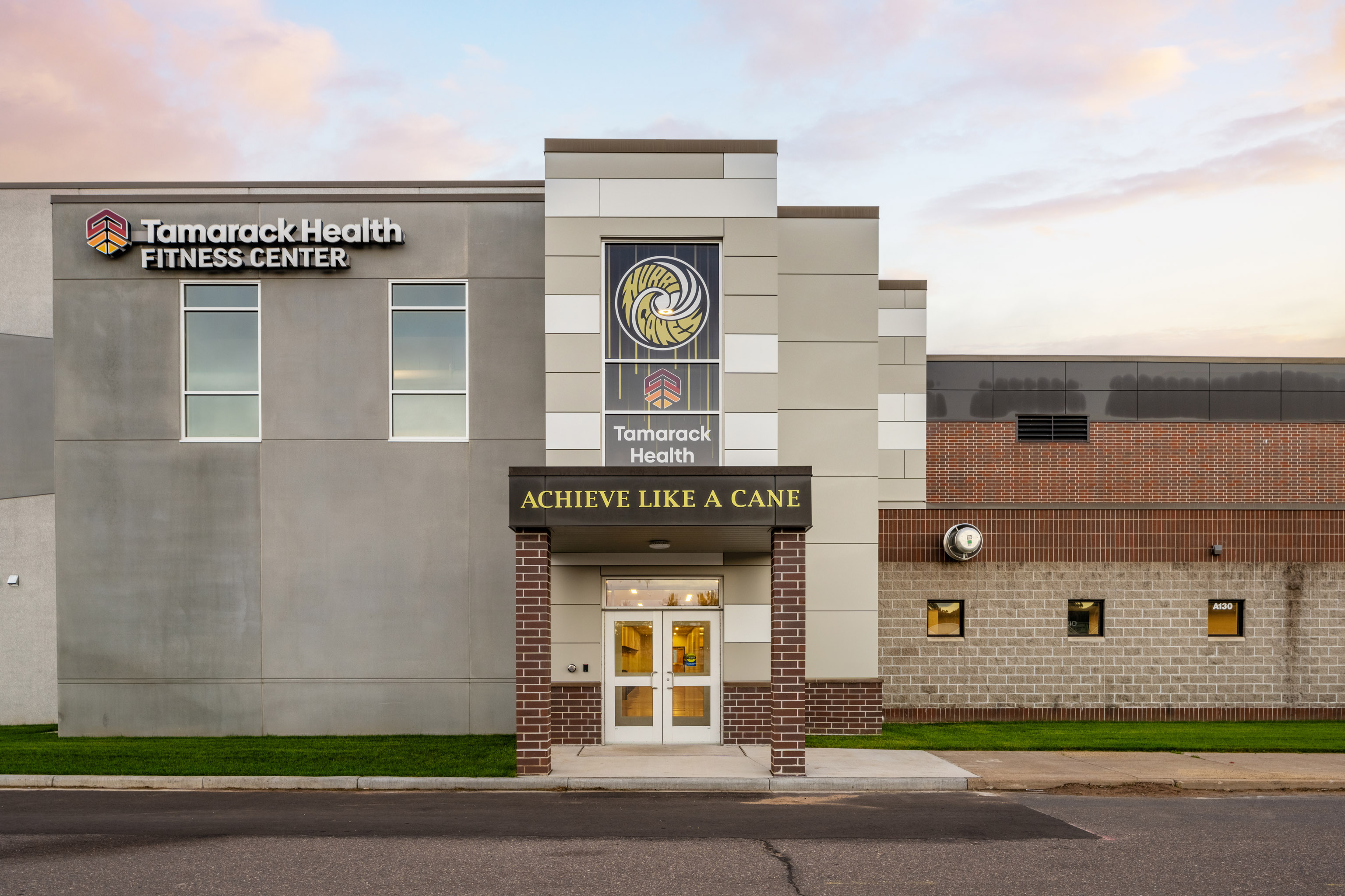 Front entrance of the Tamarack Health Fitness Center, featuring a sign that reads “ACHIEVE LIKE A CANE” above the doors, and the Tamarack Health logo and name displayed on the building.