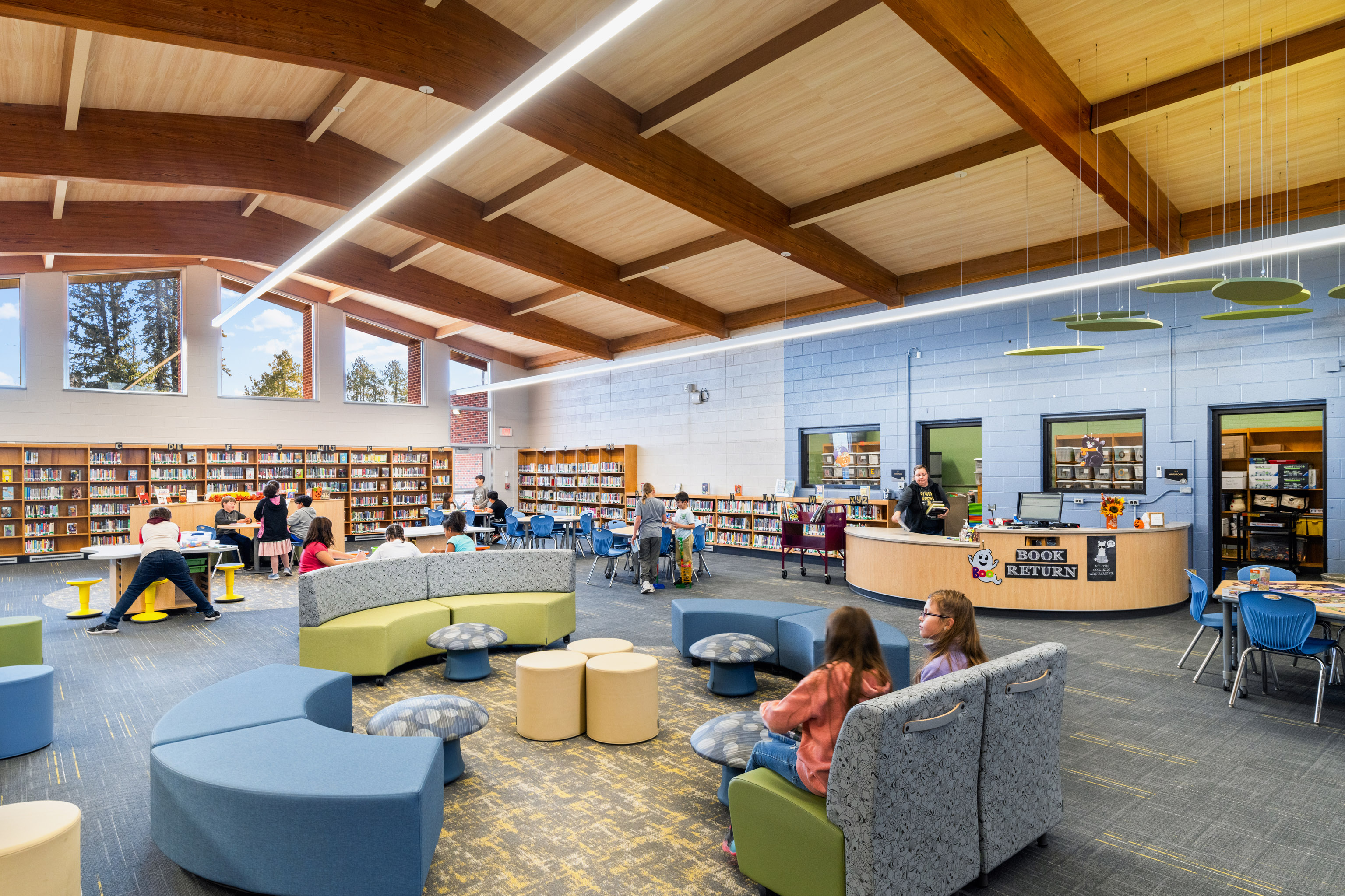A spacious, modern library with large windows, wooden ceiling beams, bookshelves along the walls, a circular seating area, and several people reading or talking. The book return desk is visible on the right.