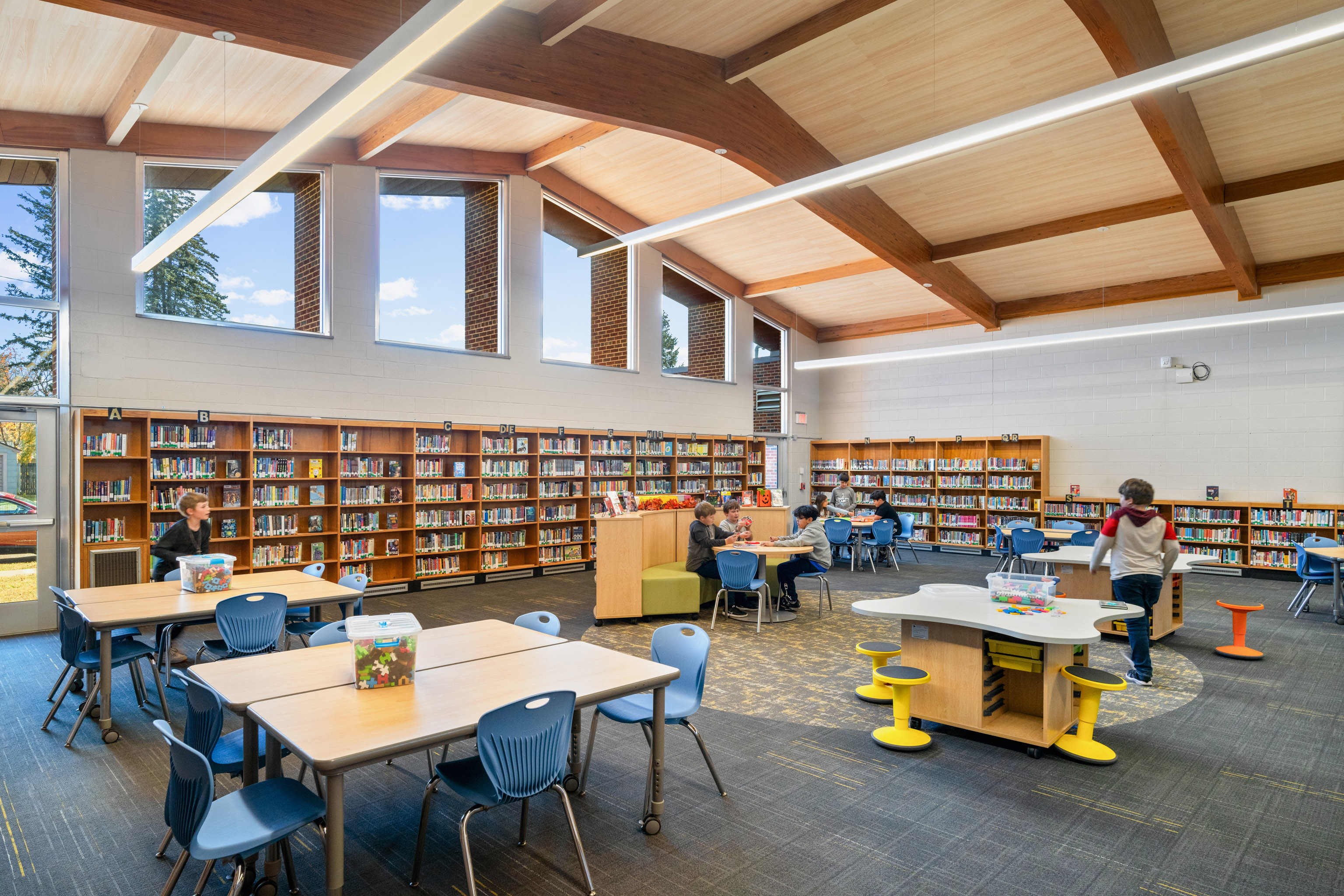 A bright, modern library with large windows, wooden beams, bookshelves lining the walls, tables with blue chairs, and a few children reading or playing at different activity stations.