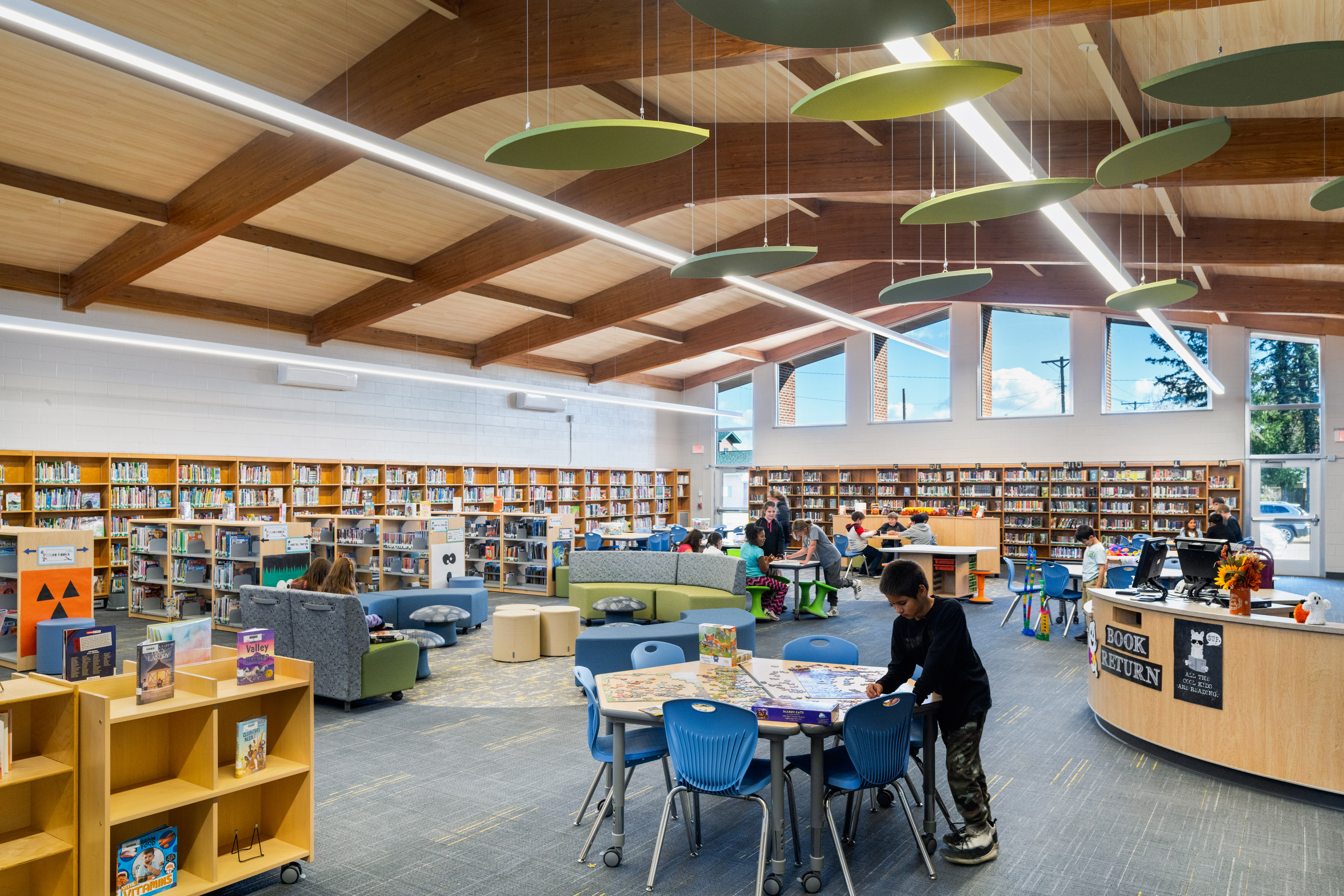 A modern library with large windows, wooden beams, bookshelves along the walls, and round tables. Children are reading and playing at the tables, while others browse books around the bright, spacious room.