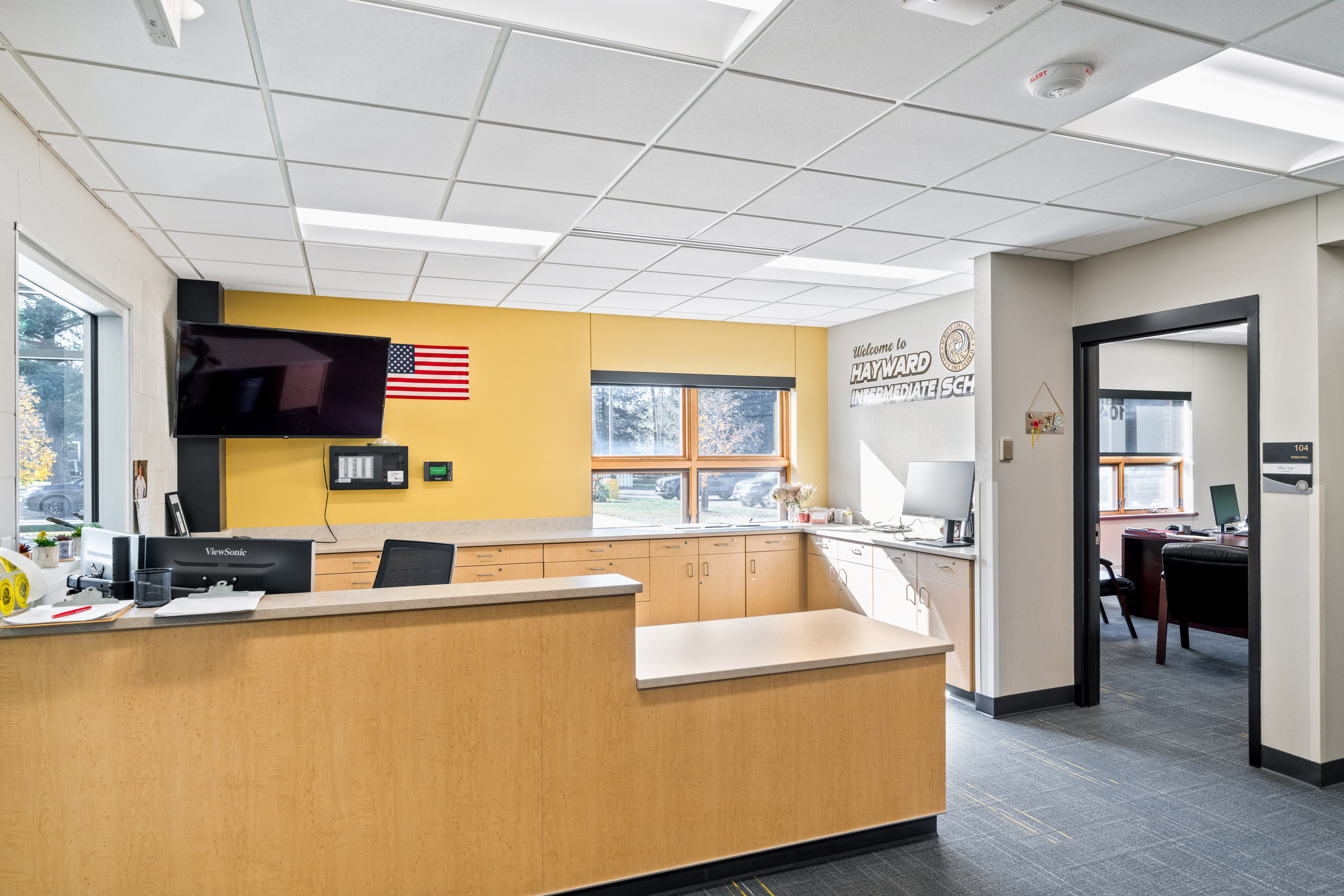 A bright school office with a reception desk, computer, and TV on the wall. An American flag and a "Welcome to Hayward Intermediate School" sign are displayed. Sunlight streams through windows behind the desk.