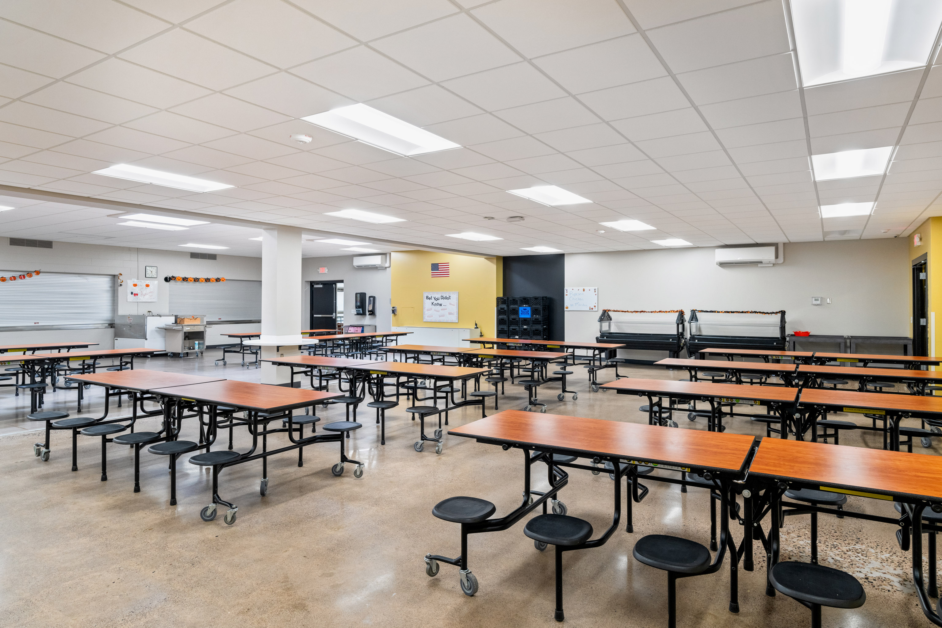 A clean, brightly lit school cafeteria with empty wooden tables and attached black stools. The room has a tiled ceiling, white walls, and posters on the walls. No people are present.