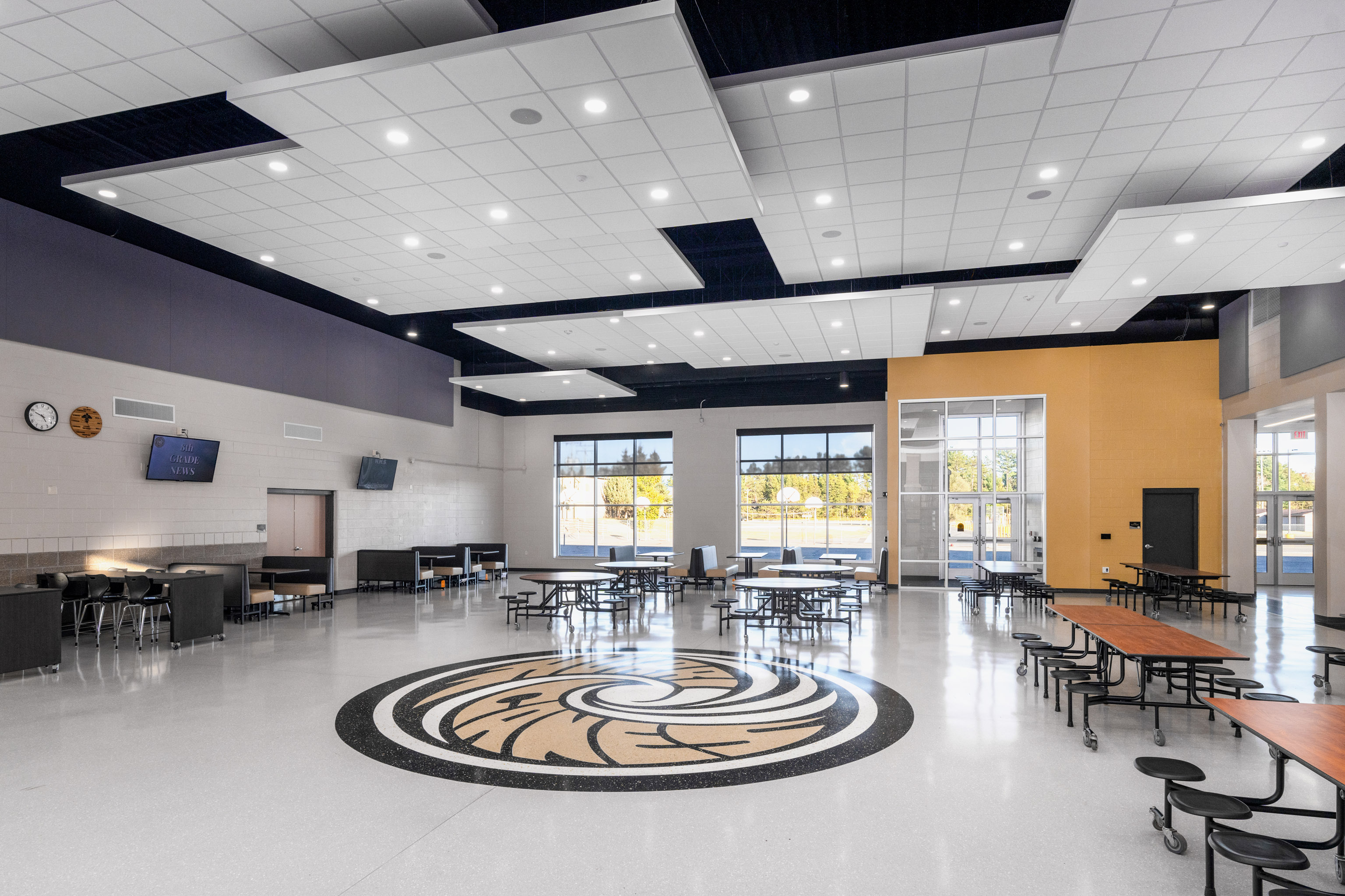 A spacious, modern school cafeteria with round and rectangular tables, a decorative floor design, large windows letting in natural light, and a high ceiling with geometric panels.