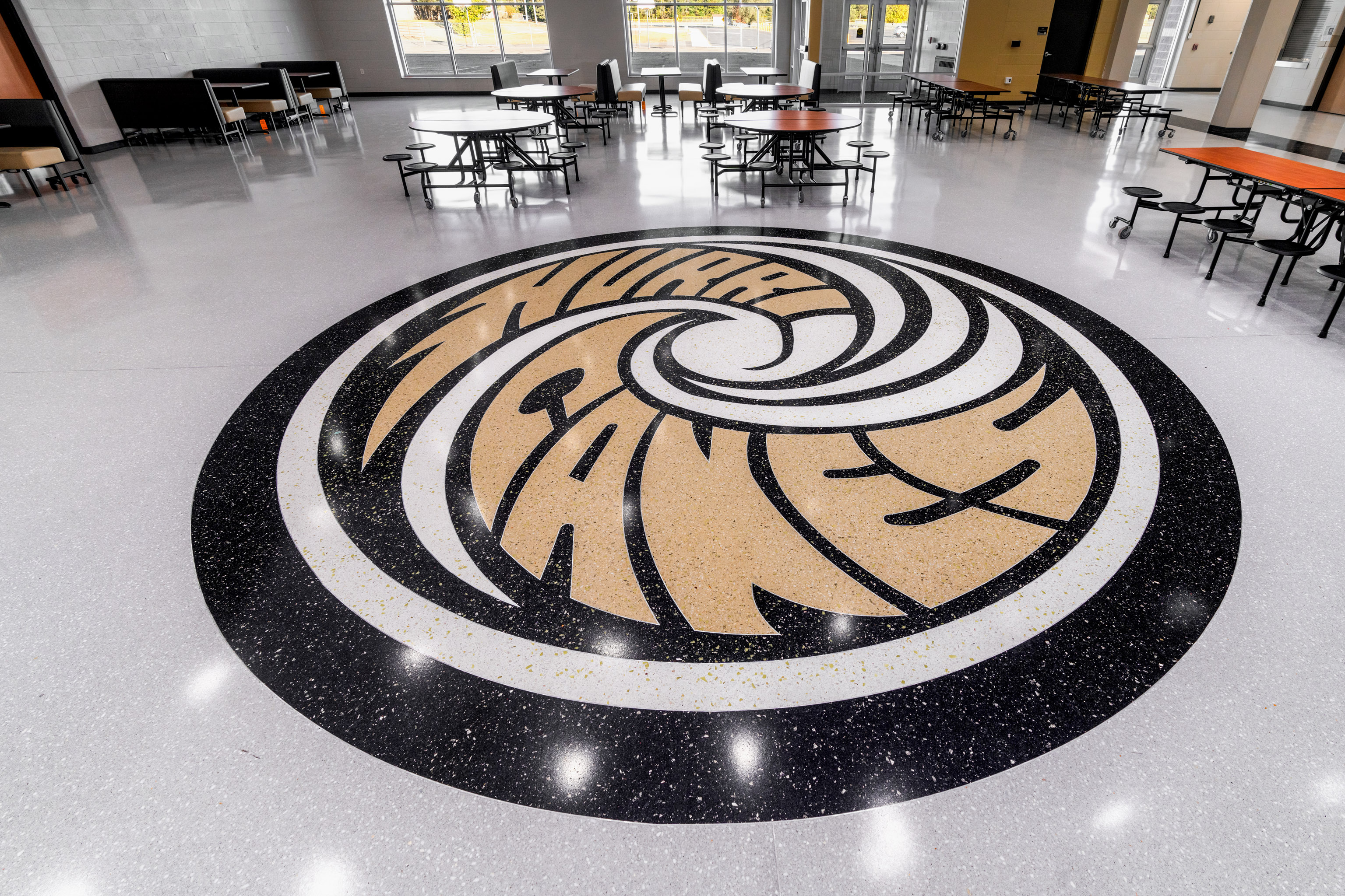 A large, circular school logo with the words "Manor Canes" is displayed on the floor of a spacious, empty cafeteria with tables and chairs around the perimeter.