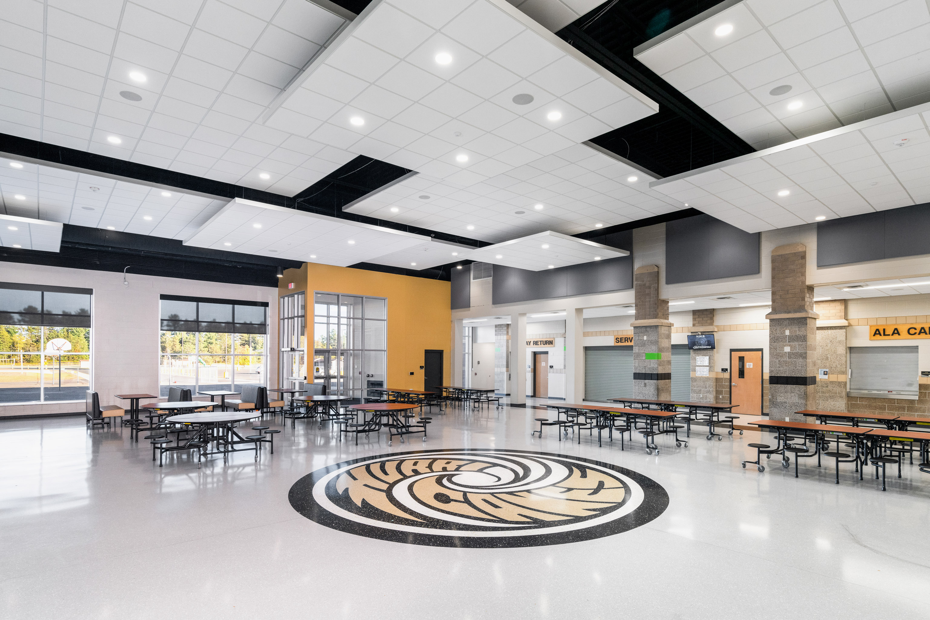 A spacious, modern school cafeteria with large windows, yellow and gray walls, long tables with benches, and a circular school logo design on the shiny floor in the center of the room.