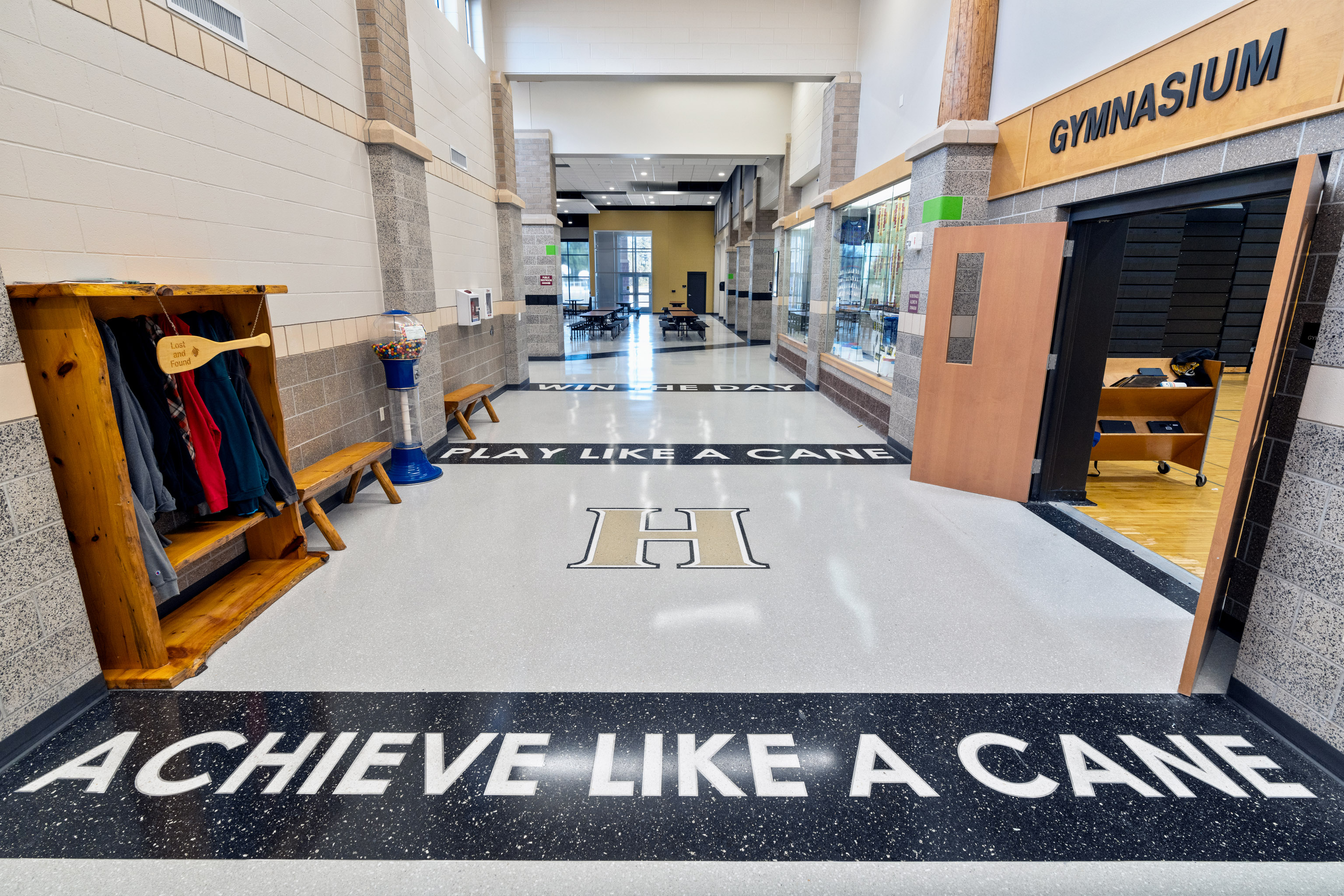 A bright school hallway with a coat rack, gym doors labeled "GYMNASIUM," and motivational phrases on the floor: "ACHIEVE LIKE A CANE," "PLAY LIKE A CANE," and "WIN THE DAY.
