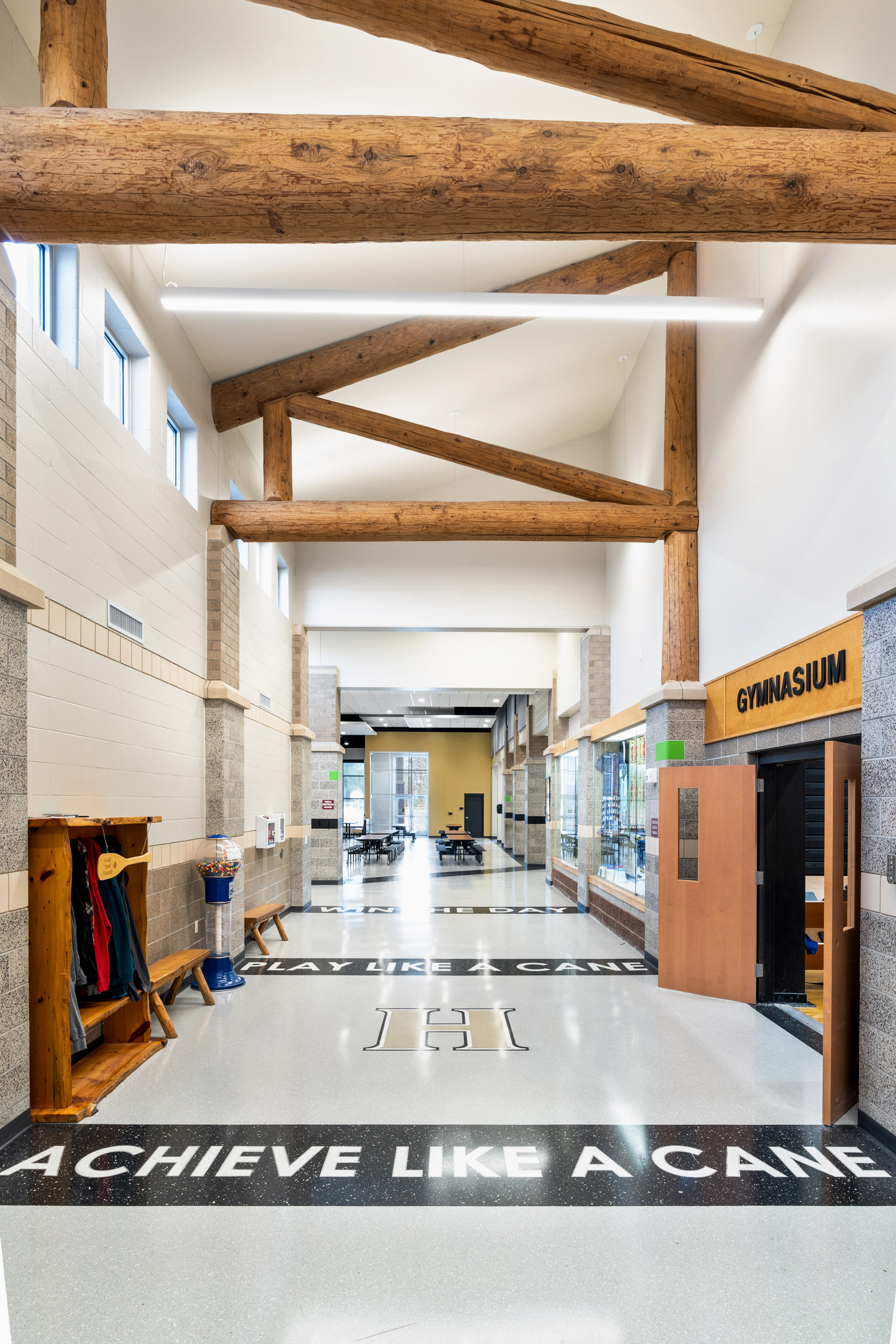 A bright school hallway with large wooden beams on the ceiling, tall windows, a wooden bench, trophy cases, and a door labeled "Gymnasium." The floor reads "ACHIEVE LIKE A CANE" in bold letters.