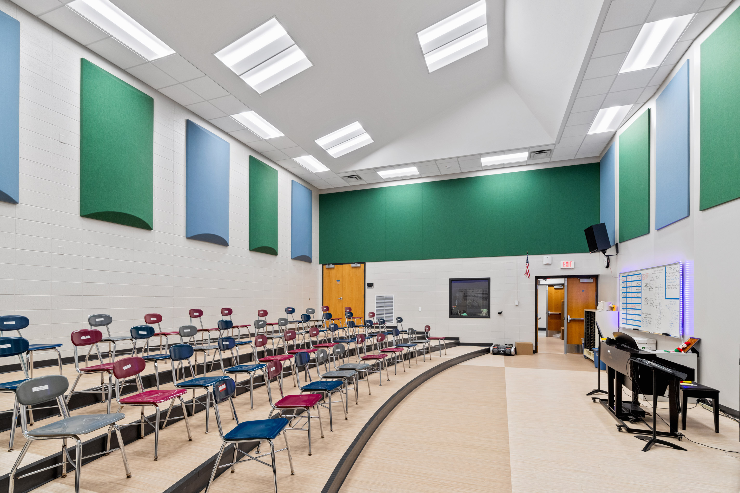A bright, modern music classroom with rows of colorful chairs in a semi-circle, green and blue acoustic panels on the walls, a piano, music stands, and a whiteboard near the front. The room has high ceilings and overhead lights.