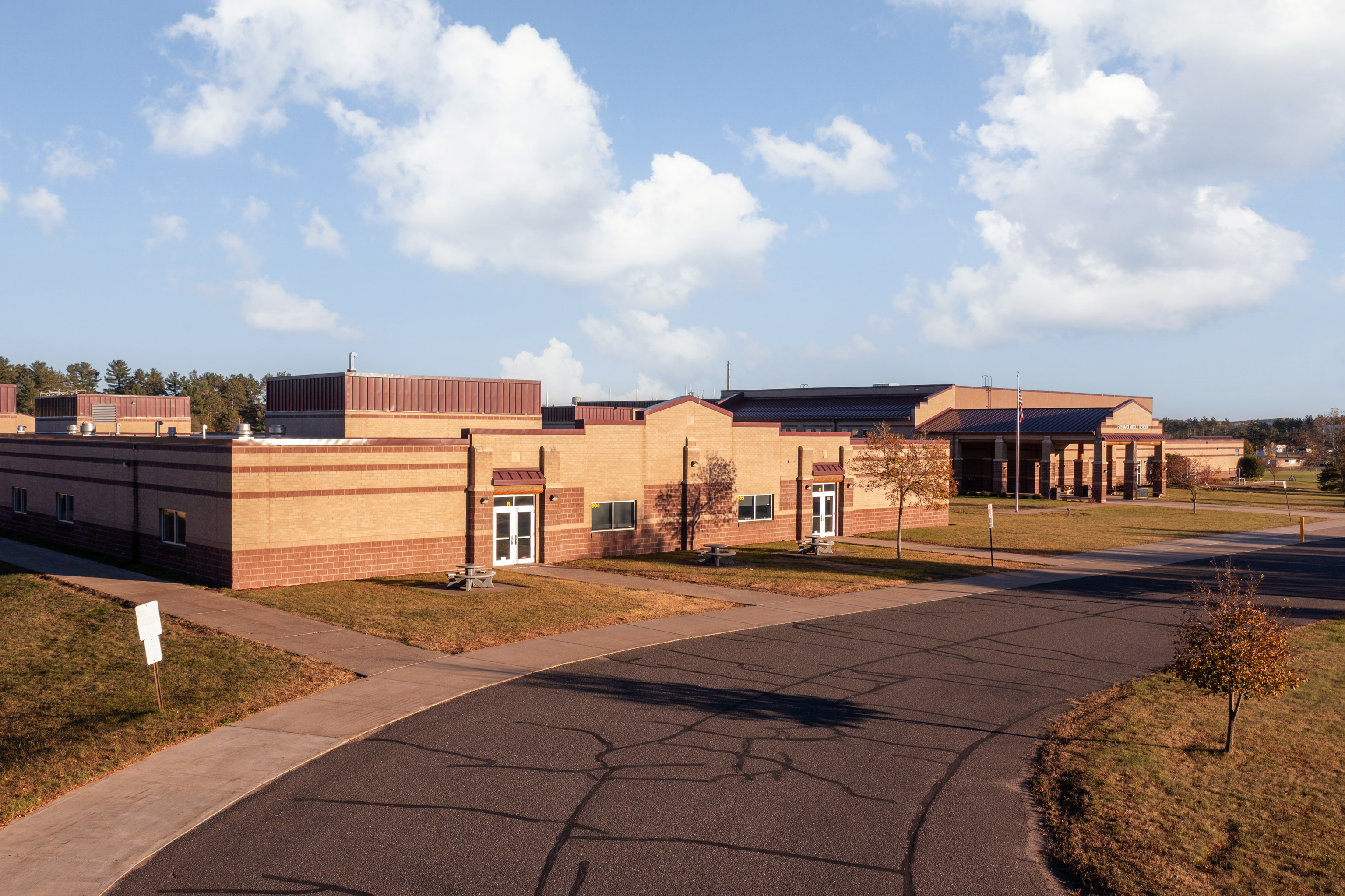 A large, modern brick school building sits under a blue sky with scattered clouds, surrounded by paved roads, sidewalks, grass, and a few small trees.