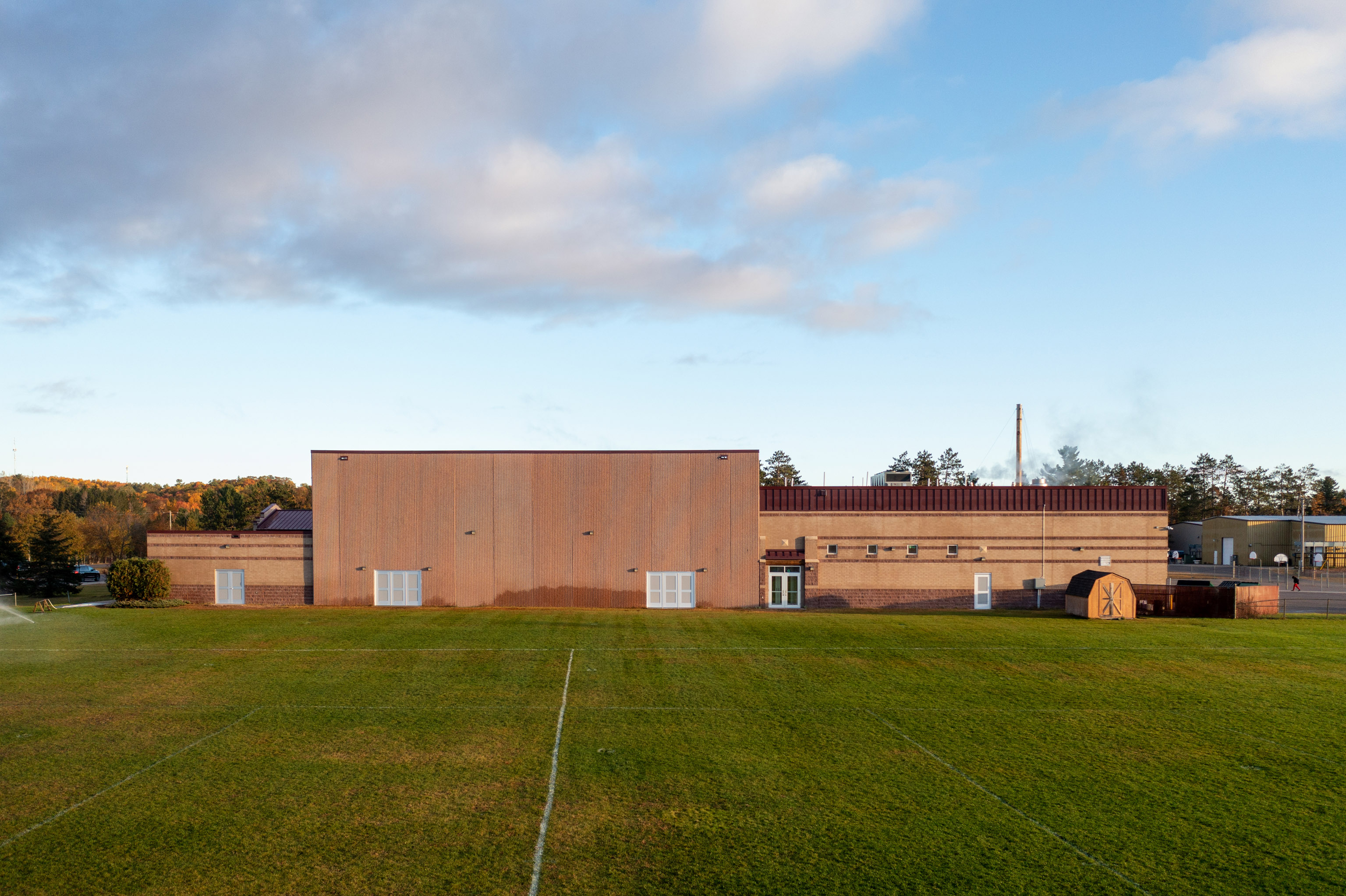 A brick school building with several white doors and windows stands behind a large grassy sports field under a partly cloudy sky. Trees are visible in the background.