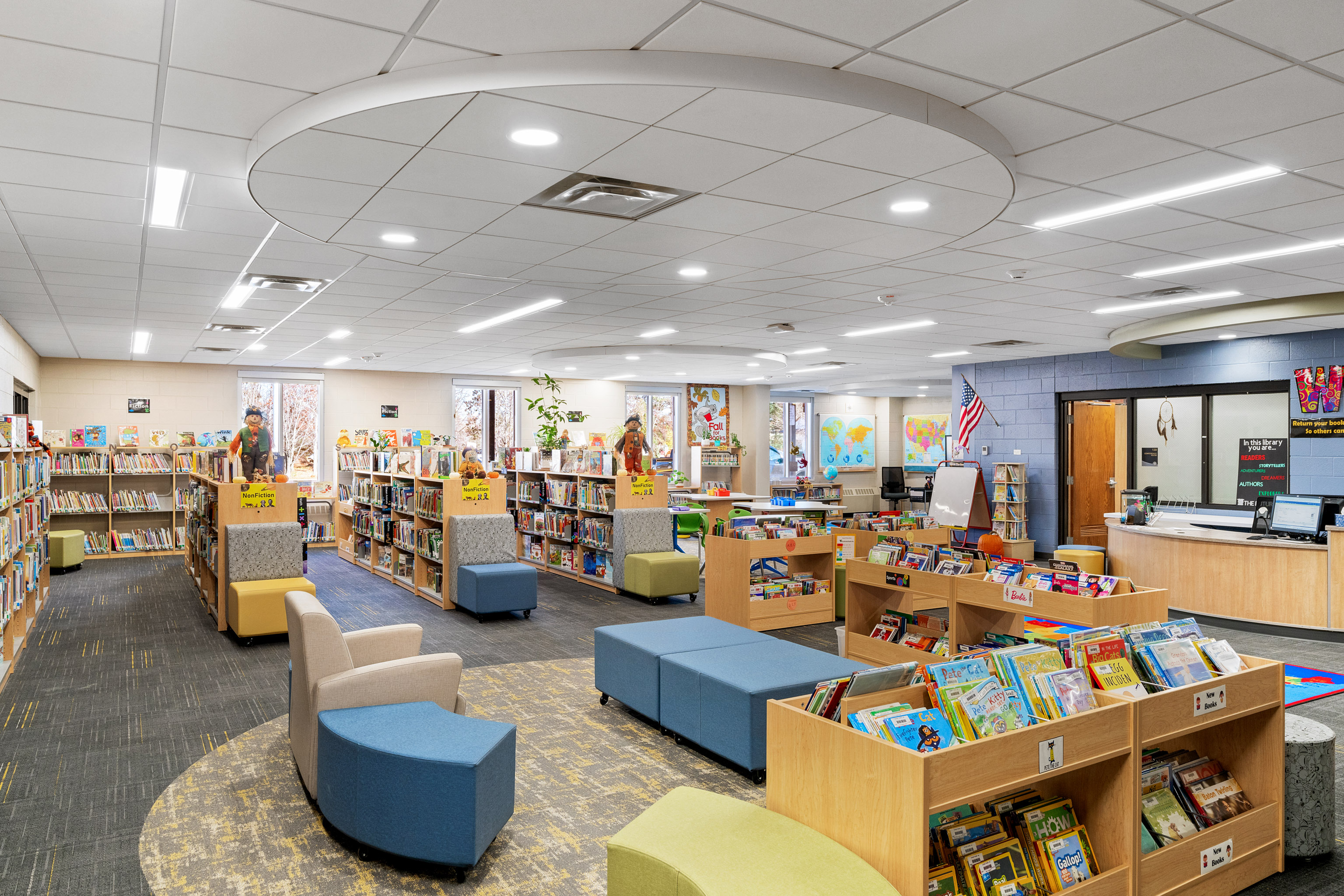 A spacious, brightly lit library with bookshelves filled with children’s books, colorful seating areas, and large windows letting in natural light. There is a front desk, reading nooks, and educational decor on the walls.
