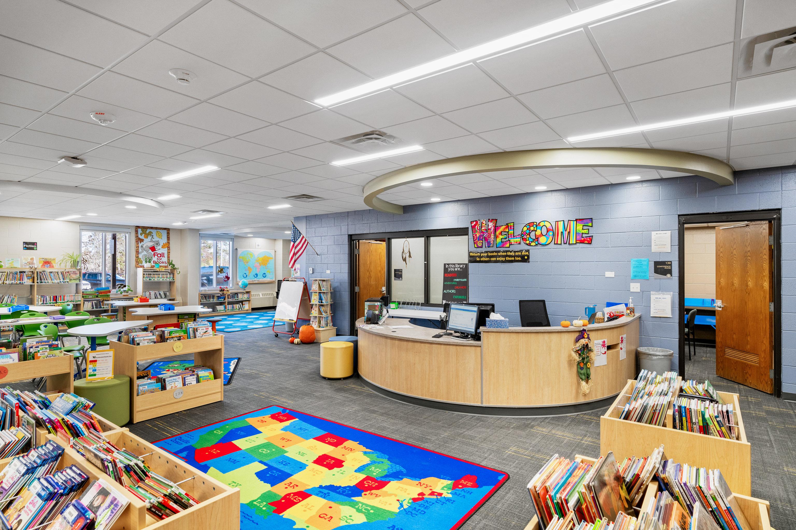 A bright, organized school library with bookshelves, colorful rugs, tables, a round welcome desk, a U.S. map rug, and a colorful “WELCOME” sign on the wall. Windows let in natural light.