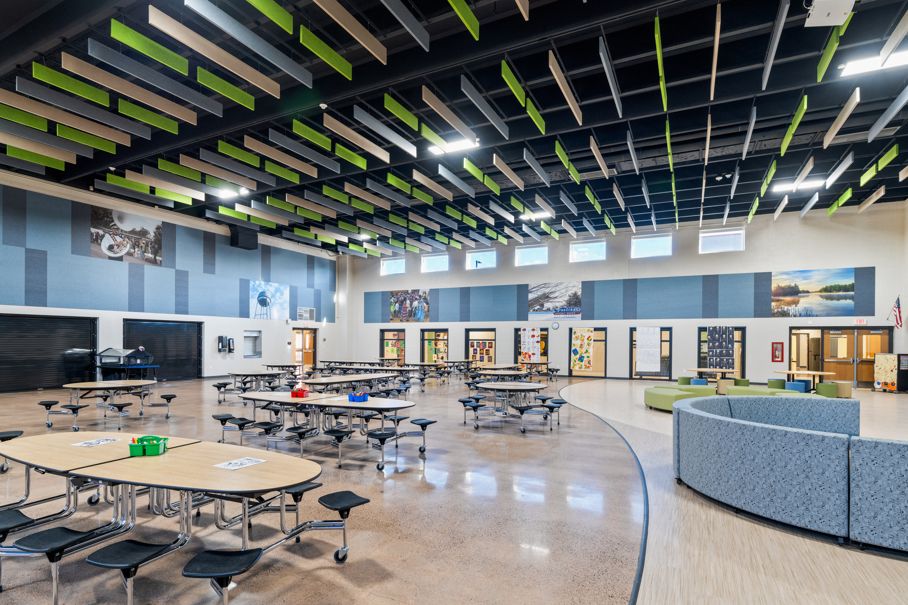 A spacious, modern school cafeteria with round tables and attached seating, a polished floor, colorful acoustic panels on the ceiling, and large windows letting in natural light. Some walls display student artwork and photos.