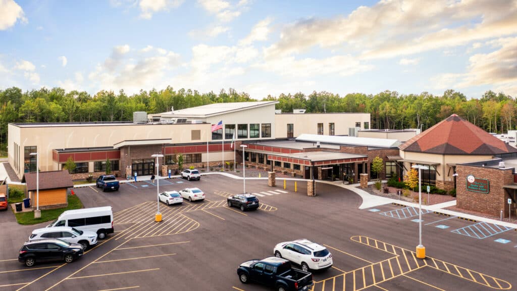A modern, multi-building healthcare facility with a brick exterior, surrounded by trees. Several cars are parked in a spacious lot with marked accessible spaces under a partly cloudy sky.