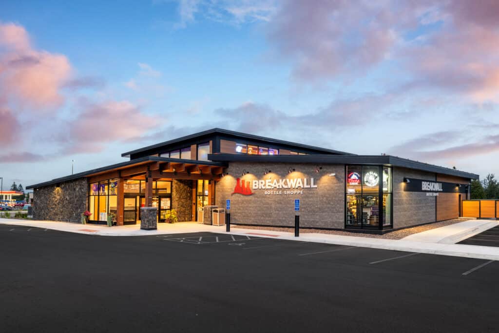 A modern, single-story brick building with large windows and a sign reading "Breakwall Bottle Shoppe" at sunset, surrounded by a spacious empty parking lot and clear sky.