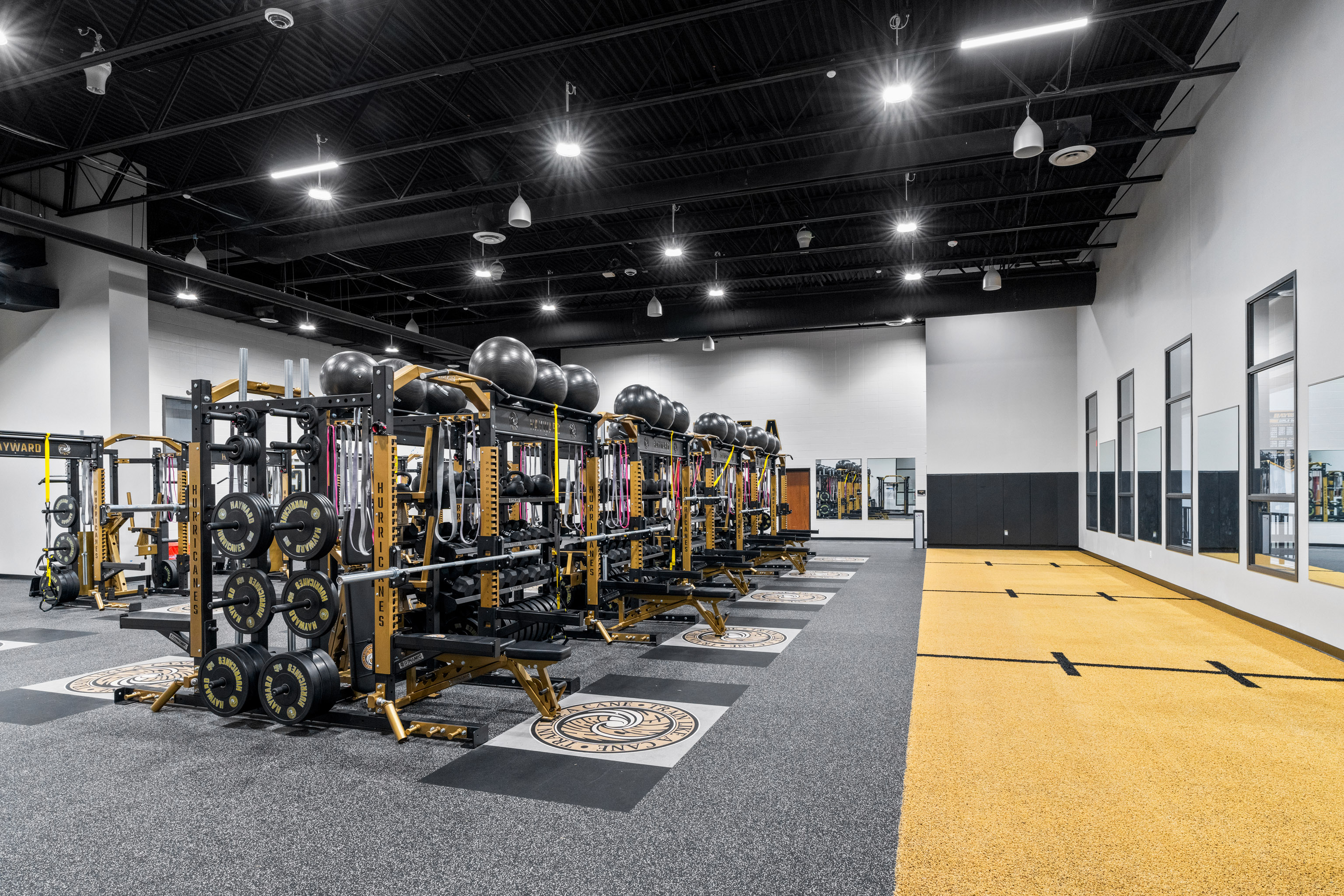 A modern gym with rows of weightlifting racks, barbells, and exercise balls on black and yellow platforms, next to a spacious area with yellow flooring and large windows on the right wall.