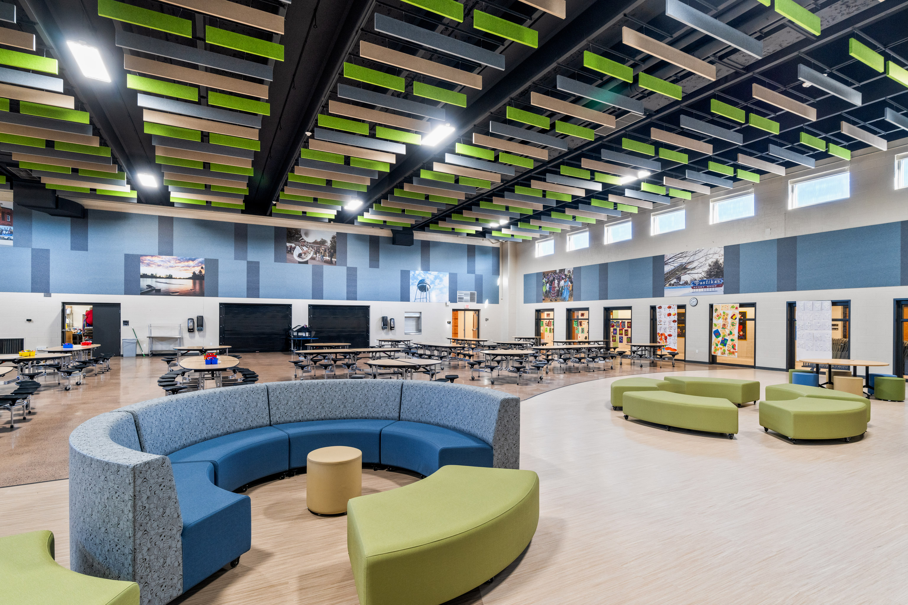 A spacious, modern school cafeteria with round and curved seating, tables with chairs, colorful ceiling panels, wall art, and large windows letting in natural light.