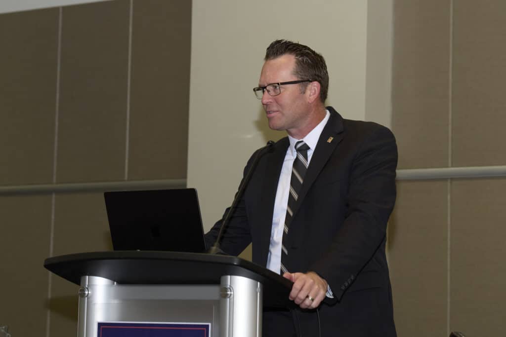 A man in a suit and tie stands at a podium with a microphone, presenting in a conference room. He is wearing glasses and looking to the side, with an open laptop on the podium. The event features Rebecca Lewis, Healthcare Architect.