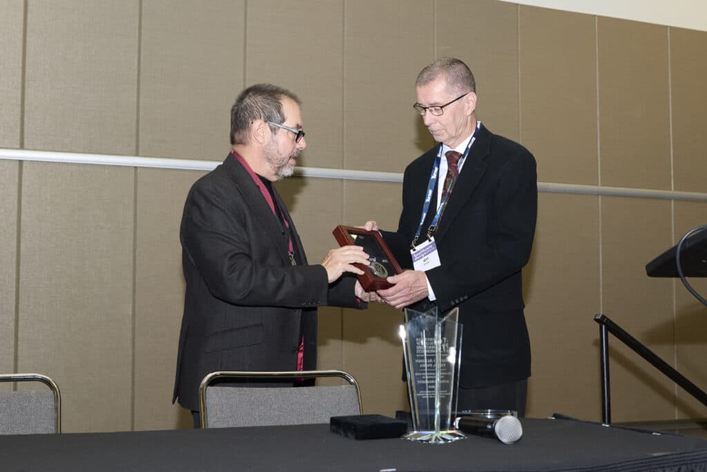 Two men in suits stand at a table, one handing an award in a wooden case to the other. A microphone and a glass trophy sit nearby. They are indoors, against a beige wall at the Rebecca Lewis Healthcare Architect event.