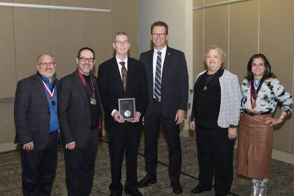 Six adults stand indoors in business attire. Rebecca Lewis, healthcare architect, is among them. The man in the center holds a plaque and two people wear medals around their necks as they smile for a photo against beige paneled walls.