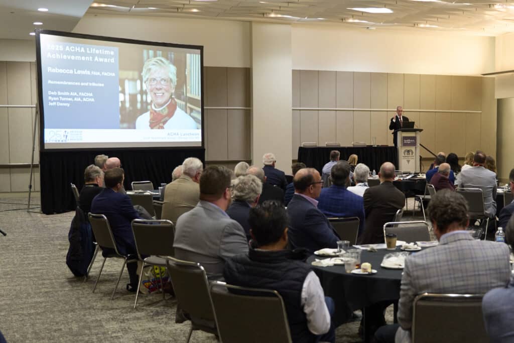 A conference room with seated attendees faces a stage, where a speaker stands at a podium. A large screen displays “2023 ACHA Lifetime Achievement Award” with a photo of Rebecca Lewis, renowned Healthcare Architect, and award details.
