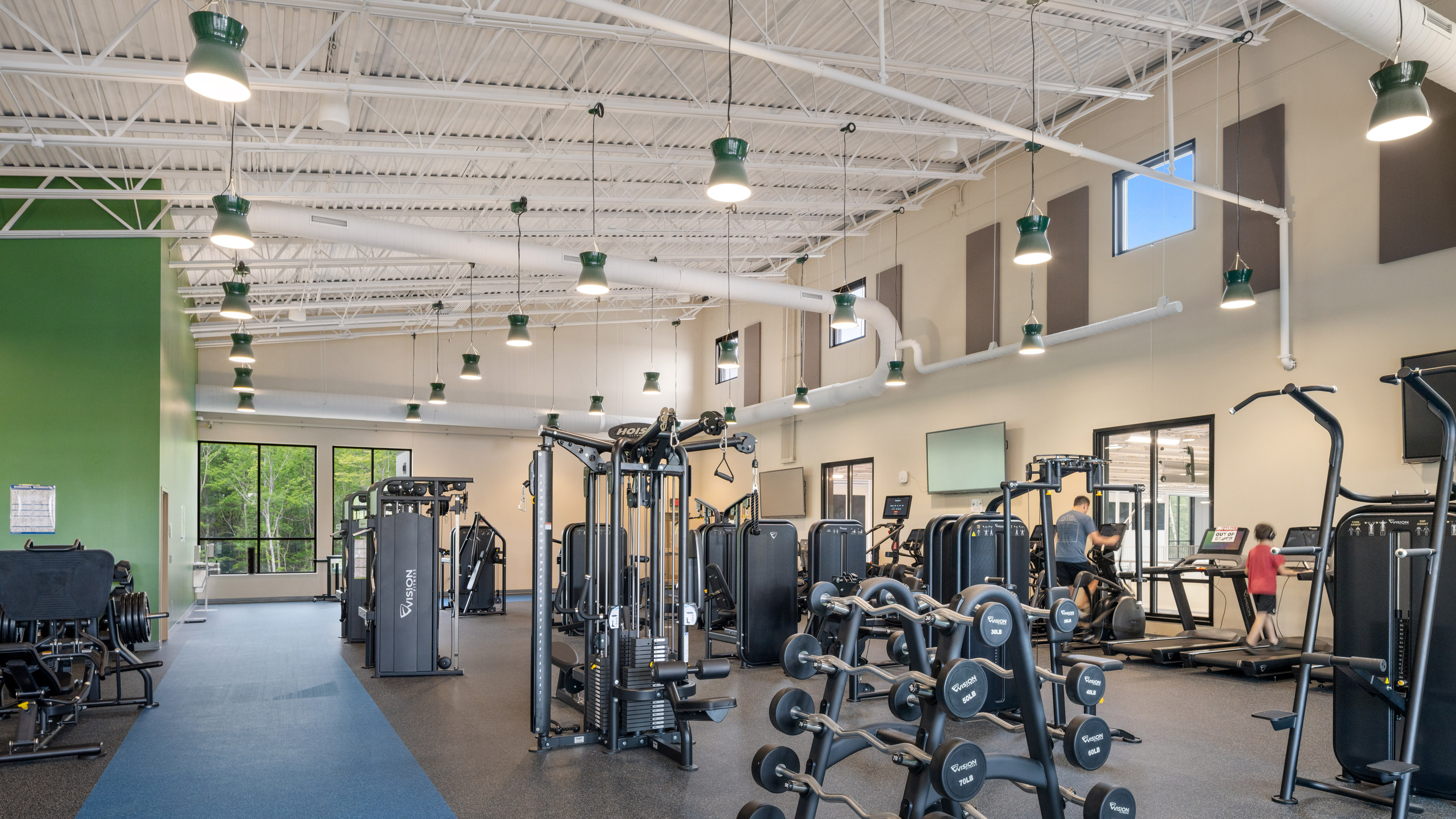 A modern gym with weight machines, free weights, treadmills, and large windows letting in natural light. Several people are working out. The space has high ceilings with exposed ducts and hanging lights.