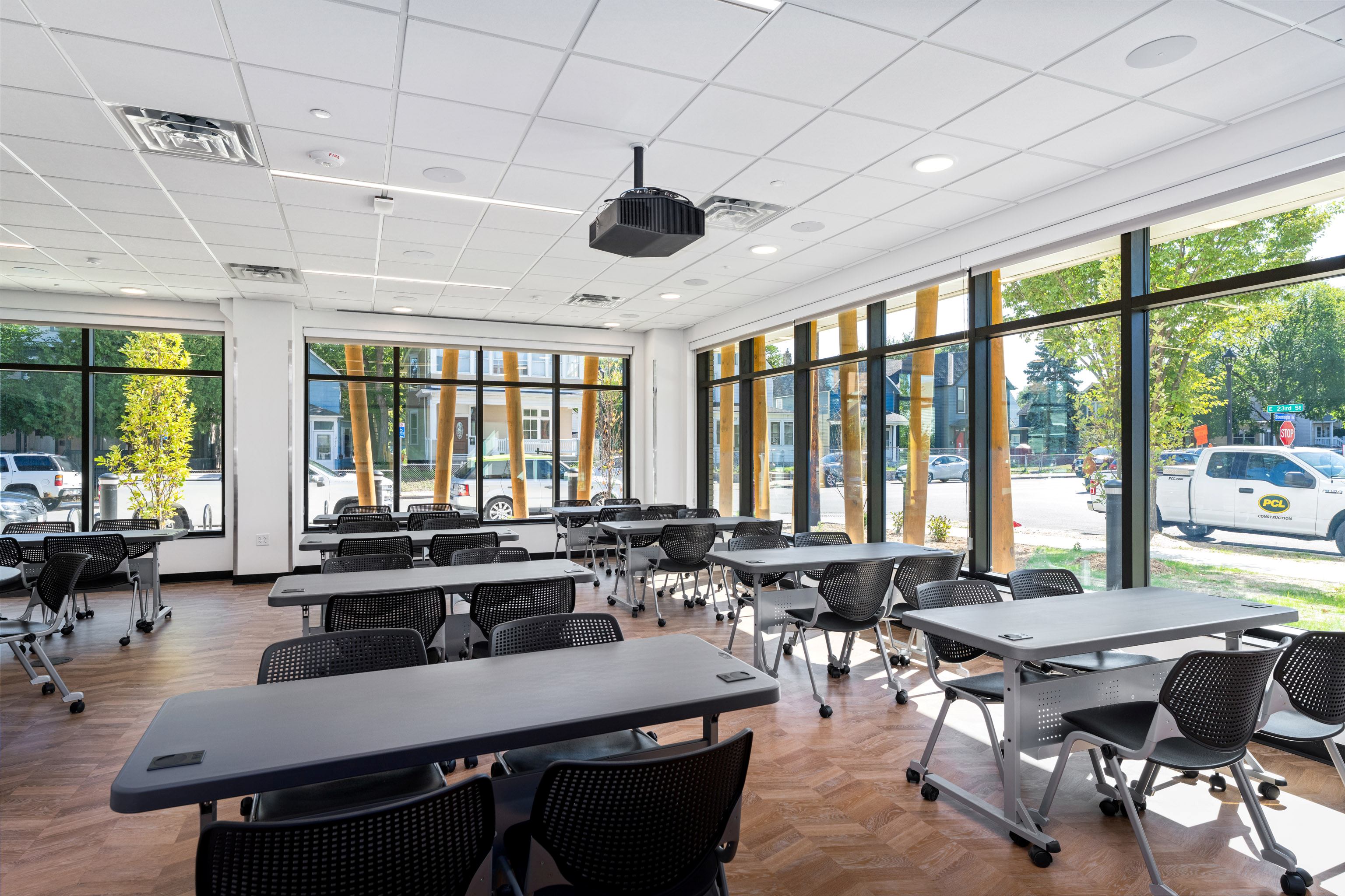 A bright, modern classroom with large windows, several black tables and chairs, a ceiling-mounted projector, and views of trees and parked cars outside.