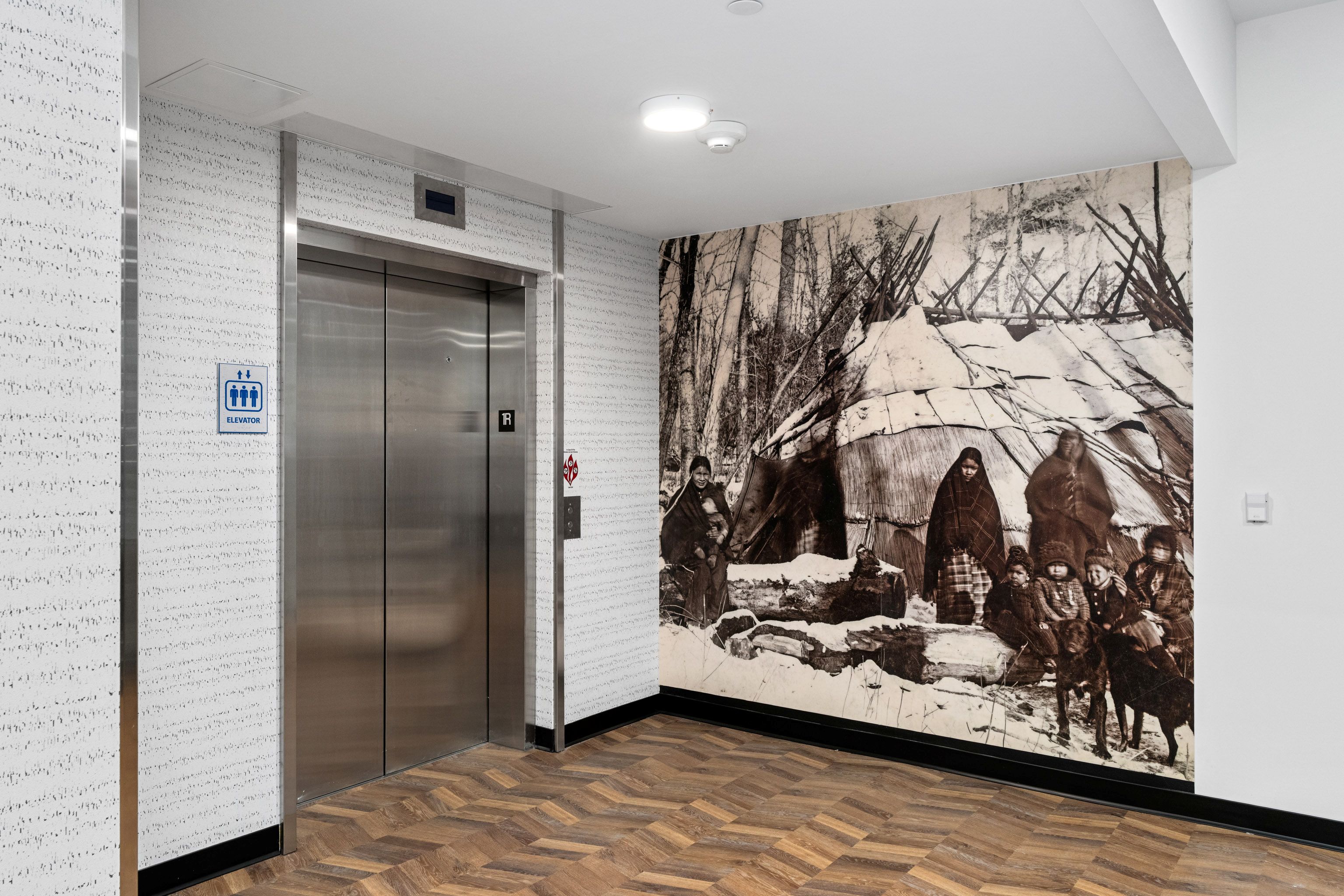 A modern building hallway with a stainless steel elevator and a large vintage photo mural of Indigenous people in front of a traditional shelter, next to herringbone-patterned wood flooring.