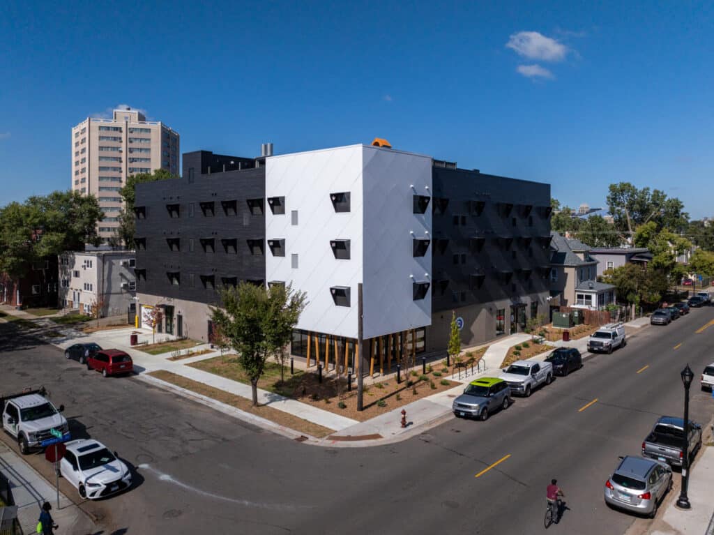 A modern, angular building with black and white exterior panels sits at a street corner. Cars are parked along the road, and a few people walk and bike nearby under a clear blue sky.