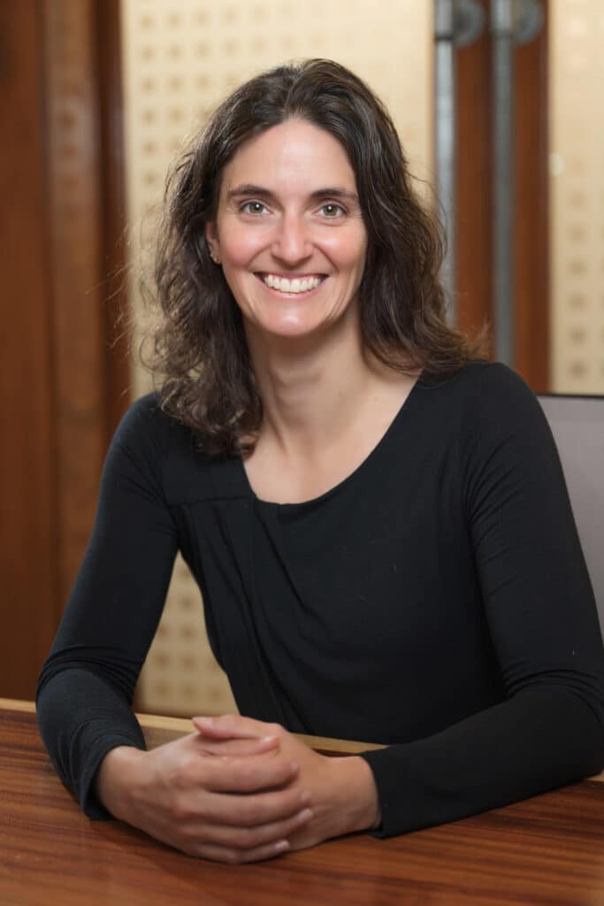 A woman with wavy dark hair, wearing a black long-sleeve top, sits at a wooden table, smiling at the camera with her hands folded. The background features wood paneling and frosted glass.