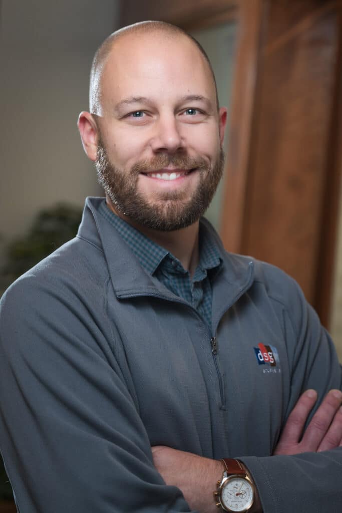 A smiling man with a beard, wearing a gray zip-up jacket and watch, stands with arms crossed in front of a blurred indoor background.