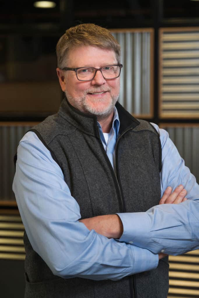 A middle-aged man with short light brown hair, glasses, and a beard is wearing a blue shirt and a dark gray vest. He is standing with his arms crossed and smiling in front of a modern, striped background.
