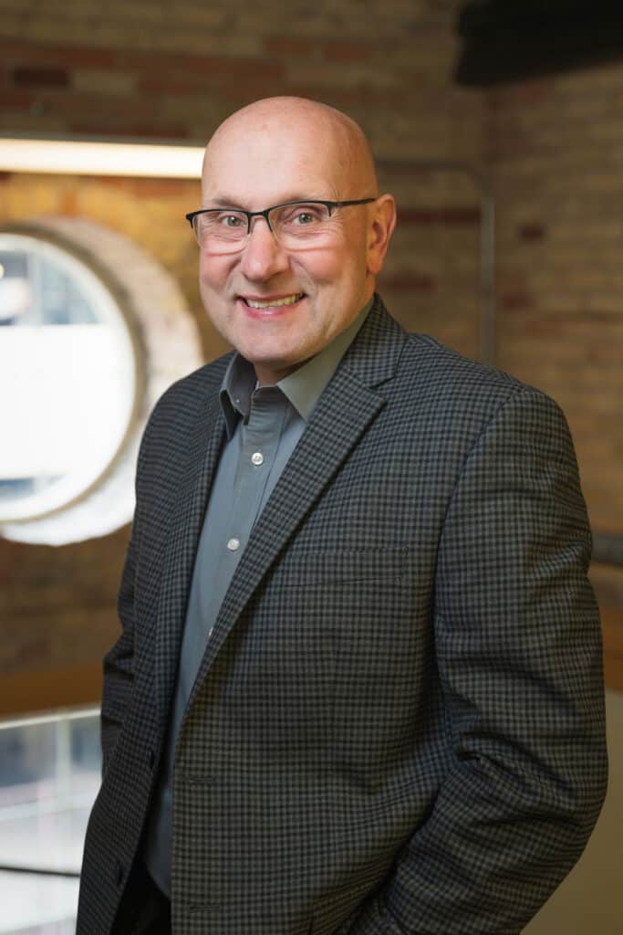 A smiling bald man wearing glasses, a gray button-up shirt, and a checked blazer stands indoors in front of a round window and a brick wall.