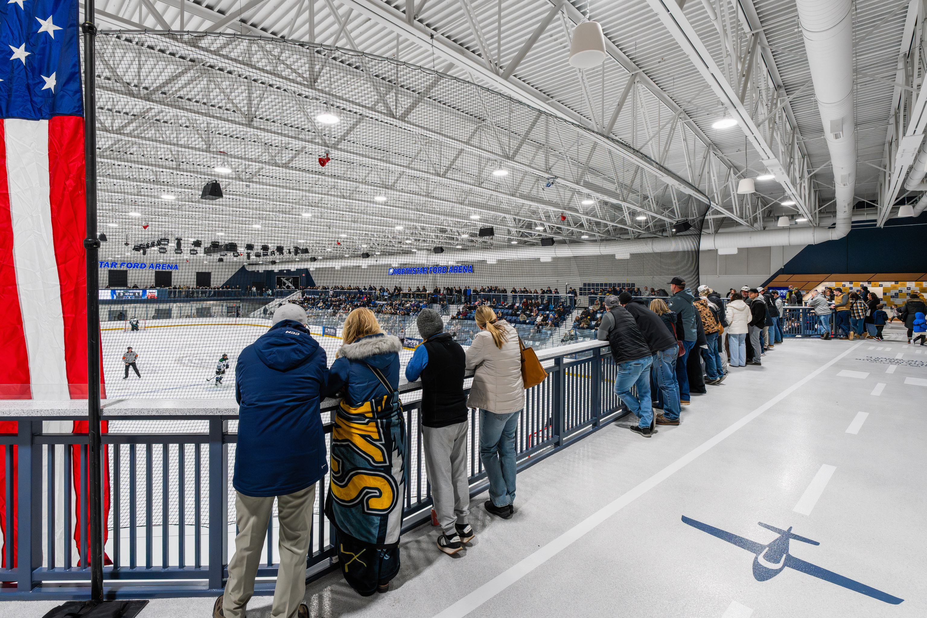 Spectators in winter clothing stand along a blue railing, overlooking an indoor ice hockey rink. The venue is brightly lit with high ceilings, and an American flag hangs on the left side of the image.